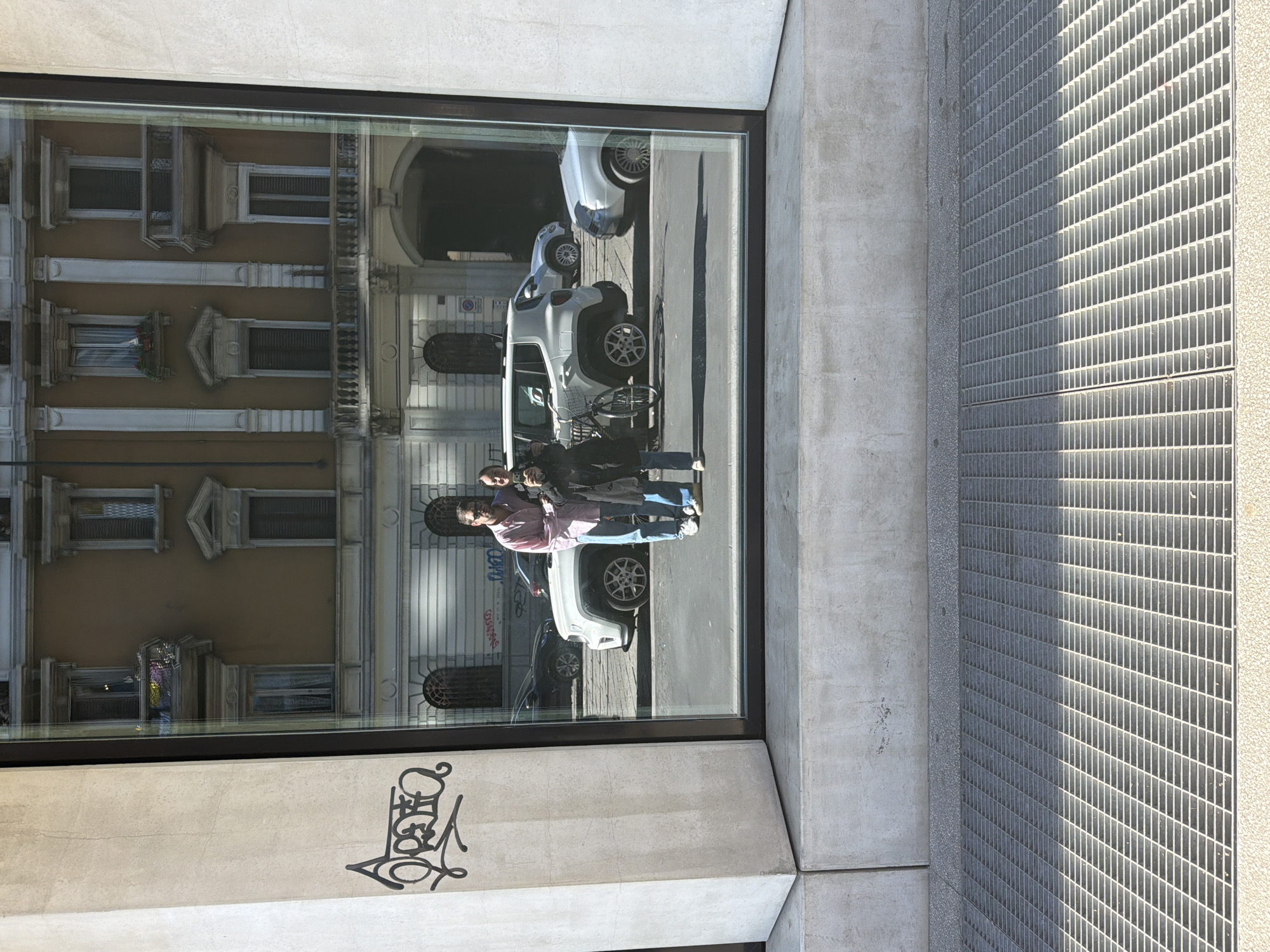 The image displays a street-level view dominated by a large reflective glass window set within a modern concrete building. The window acts as a mirror, capturing a reflection of the opposite side of the street, while the foreground shows elements of the building and ground.
In the reflection, two individuals are visible. One person, wearing a pink shirt and blue jeans, is holding a smartphone, seemingly capturing an image or video. Another person, dressed in dark clothing, stands beside them. Also reflected are two white Jeep Renegade-style SUVs parked on the street, with a bicycle positioned near the front vehicle. In the background of the reflection, an ornate, multi-story building with multiple windows, balconies, and an arched entrance typical of European architecture can be seen, consistent with the Milan, Italy location.
In the physical foreground, the concrete wall beneath the window features stylized black graffiti. To the right of the window, a large metal grate with a distinct grid pattern covers the ground.
The scene appears to take place during daytime, indicated by bright sunlight and visible shadows cast across the metal grate. The weather is sunny and clear. The main activity involves the two reflected individuals standing on what appears to be a sidewalk, with one using a phone.