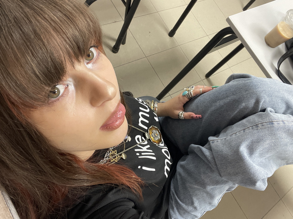 An upward-angled close-up depicts a female individual seated indoors. She has brown hair with reddish highlights and bangs, green-brown eyes, light-colored lip gloss, and white eyeliner. She wears a black long-sleeved shirt with the partial white text "i like mus" and light blue denim jeans. Numerous accessories include multiple silver-toned necklaces featuring a Star of David pendant, a heart charm, and a large ornate round pendant. Her left hand displays several rings, notably one with a large turquoise stone, and multiple bracelets. Her long, manicured nails are painted with various colors and designs, including teal/green and pink with black text. The background reveals a light-colored tiled floor, black metal chair legs, and a partial white surface, presumably a table, holding a light brown beverage cup and a clear plastic bottle. The specific location in Raanana, Israel, is not discernible from the image.