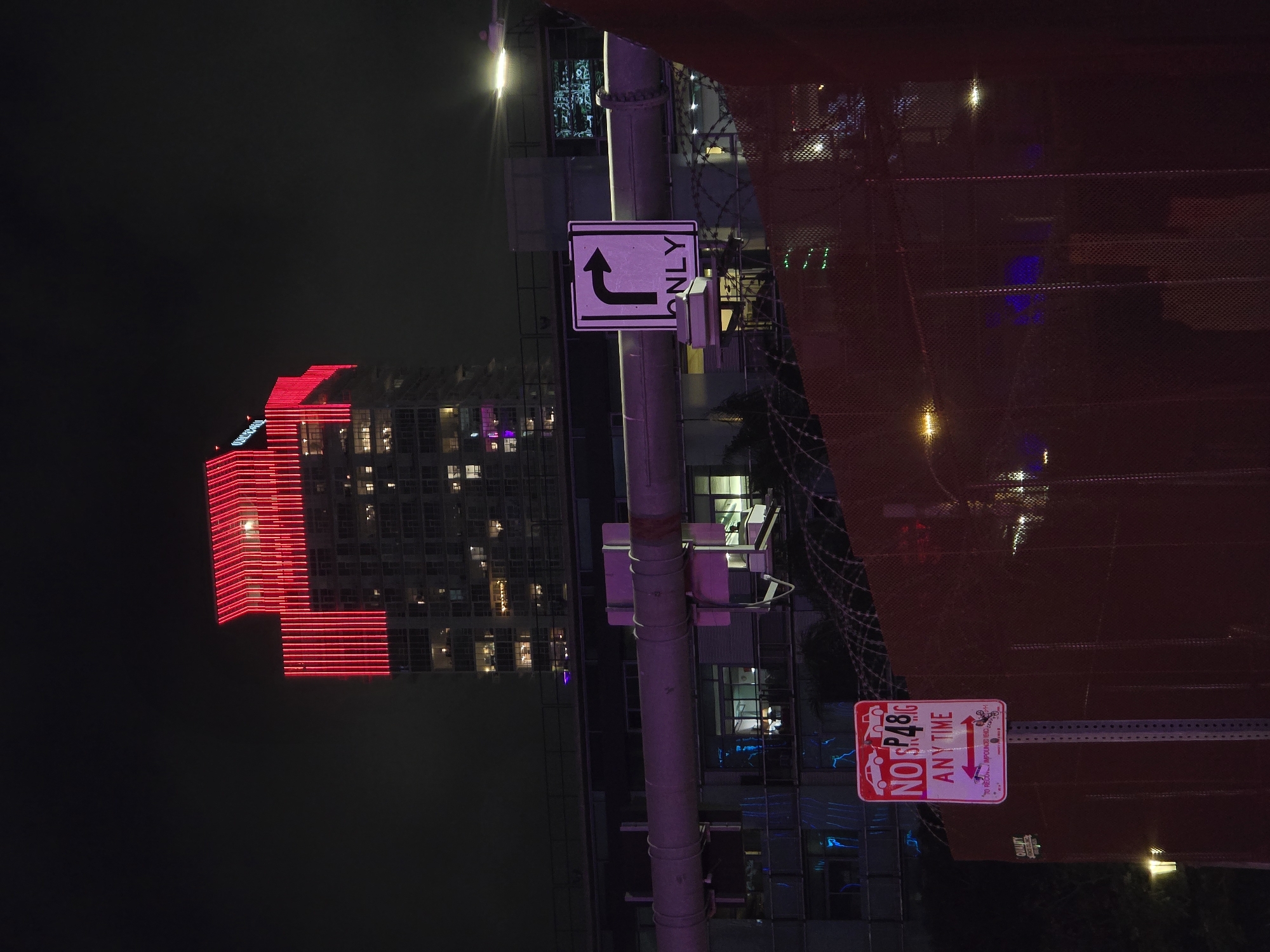 The image depicts a night scene in an urban environment, likely in Los Angeles, United States of America, characterized by a towering skyscraper illuminated with vibrant red lights. The building features a modern architectural design with distinct vertical red LED lighting accentuating its upper floors and roofline. In the foreground, a metal fence, partially obscured by a reddish-brown mesh material, stretches across the frame. Two street signs are visible on metal poles in front of the fence. One sign is white with red lettering and symbols, indicating "NO PARKING ANY TIME" with arrows showing towing information. The other sign is a white square with a black curved arrow pointing right, accompanied by the word "ONLY" in black text below it, signifying a turn restriction. A large, cylindrical pipe, possibly part of an industrial structure or elevated walkway, runs horizontally in the mid-ground, reflecting pink and purple light. The background shows more buildings with illuminated windows, suggesting a densely populated urban area. The overall atmosphere is dark, with artificial lighting dominating the scene.
