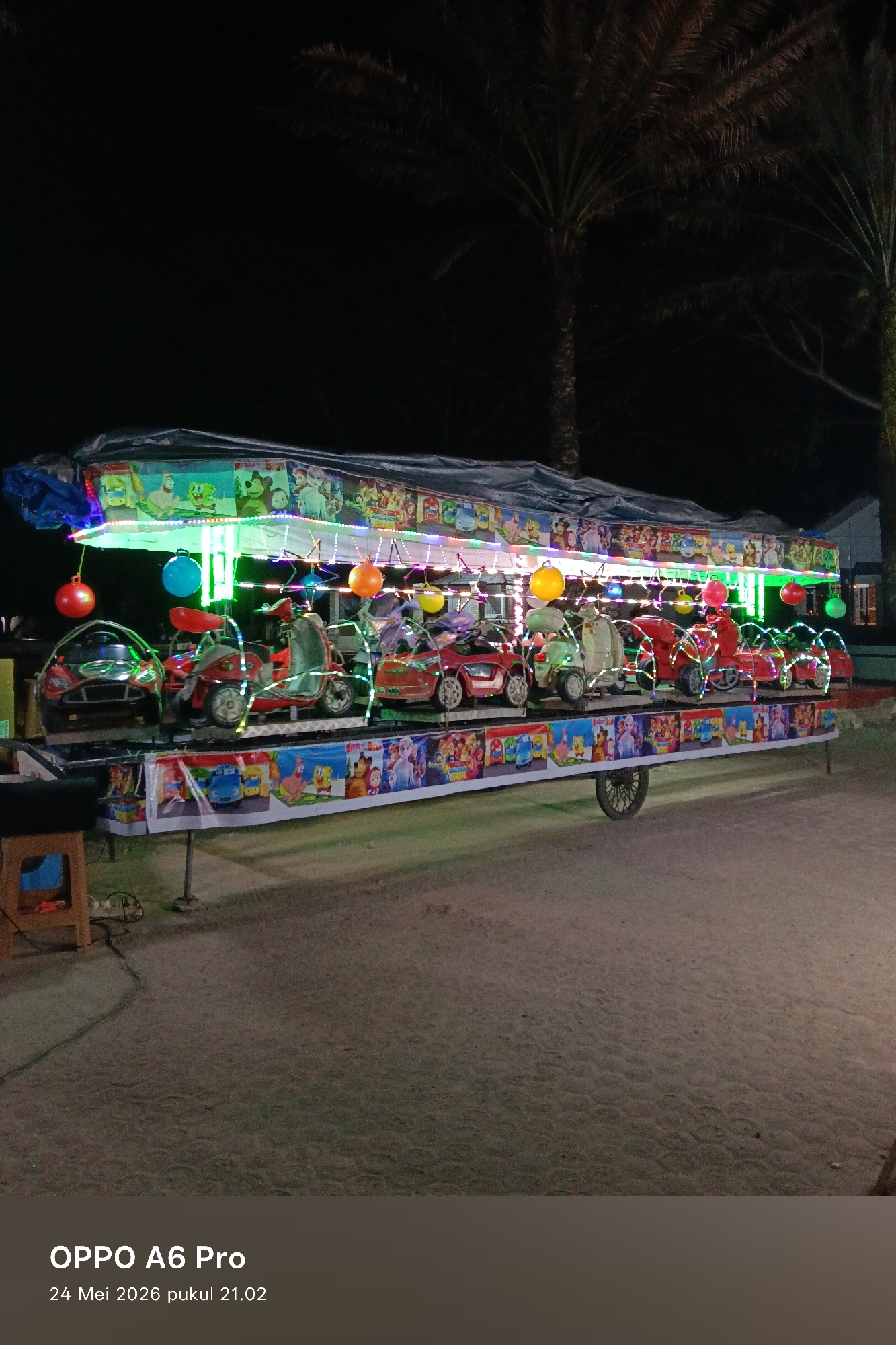 The image depicts a children's amusement ride at night. The location appears to be outdoors in a public area, possibly a fairground or park, within the city of Unknown, Unknown.
The ride consists of a platform on wheels featuring a variety of vehicles, including cars and scooters, each decorated with lights. The upper portion of the ride is adorned with cartoon characters.
A small wooden stool is visible near the front of the ride. The ground surface appears to be paved. Tall trees are visible in the background, suggesting an open-air environment.