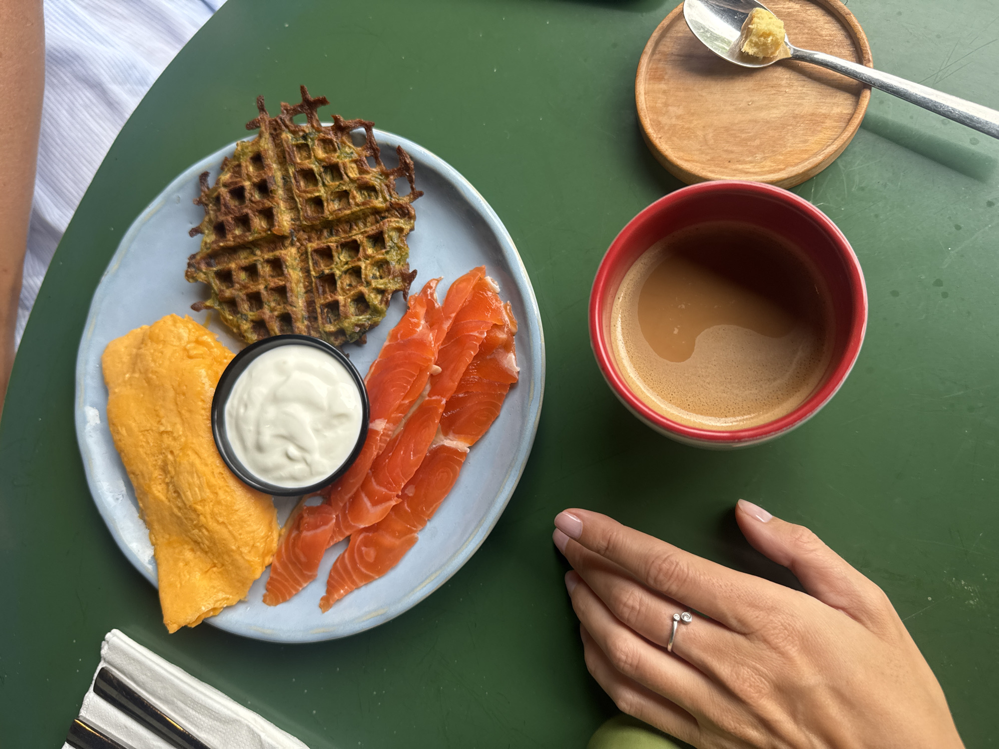 An overhead shot shows a meal on a green table. On a pale blue plate, there is a large piece of orange-yellow omelet, several slices of smoked salmon, a small black bowl filled with white sauce, and a savory waffle. A red mug of coffee with foam sits to the right of the plate. Next to the coffee mug is a wooden coaster with a spoon holding a piece of yellow food. A person's hand with a silver ring is visible on the left side of the frame, resting on the table. The background shows a glimpse of white and blue striped fabric. The scene is set in the city of Mengwi, Indonesia.