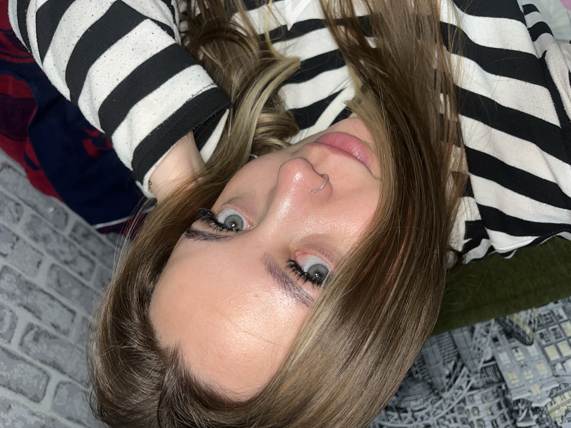 A close-up, slightly rotated image captures a woman from a low angle. Her eyes are light-colored and framed by dark eyelashes, possibly enhanced. She has a nose ring. Her hair is light brown and blonde. She is wearing a black and white striped long-sleeved shirt. In the background, there is a brick wall and a bedding with a cityscape pattern on it. There is not enough information to accurately identify the location as Novopokrovskaya, Russia.