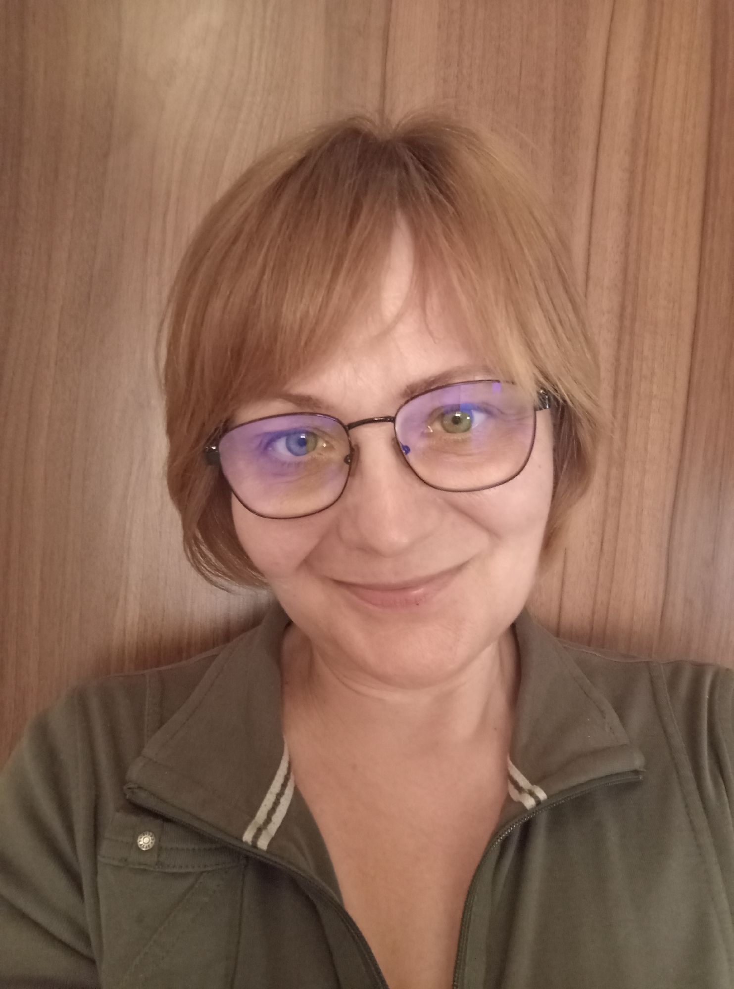 A medium shot selfie is captured from chest up, featuring a woman with shoulder-length auburn hair and fair skin, wearing clear-rimmed glasses. Her eyes are blue and green with reflections in the lenses. She is dressed in a dark olive green zip-up jacket with white stripes on the collar. The background is a plain wooden wall. The image was taken indoors, likely in a residential setting within the city of Brno, Czechia.