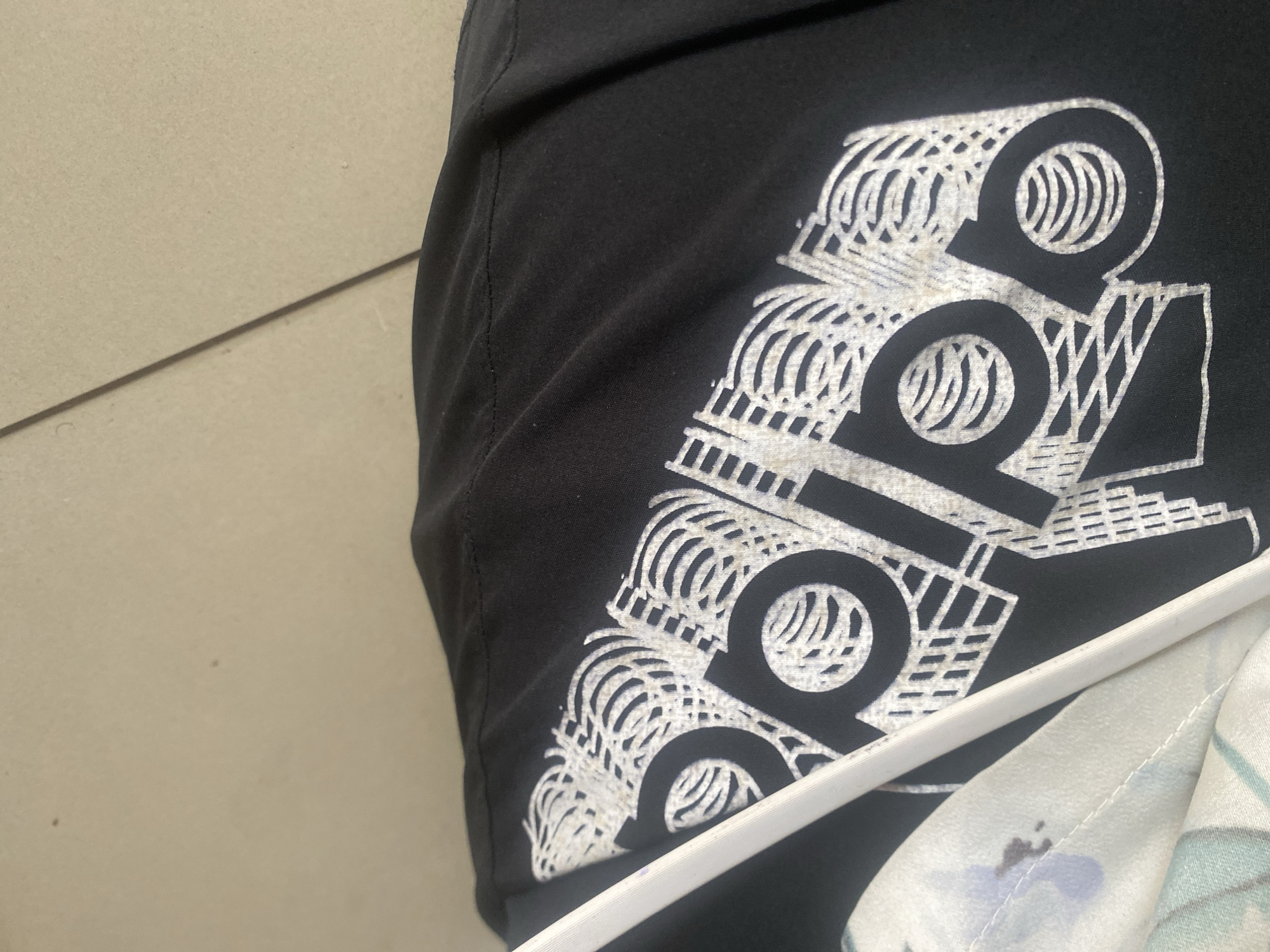 The image shows a close-up of a black t-shirt with a white Adidas logo printed on it. The t-shirt is laid flat, and part of another garment with a floral pattern is visible underneath it. A white hanger is also visible, positioned diagonally across the t-shirt, obscuring part of the logo. The background appears to be a light-colored, textured surface, possibly a wall or floor. The lighting is somewhat dim, creating shadows on the t-shirt. The scene is photographed from a slightly elevated angle, looking down at the garments. There are no people visible in the image. The location of Wauro Leggal, Nigeria is not evident from the visual information provided in the image.