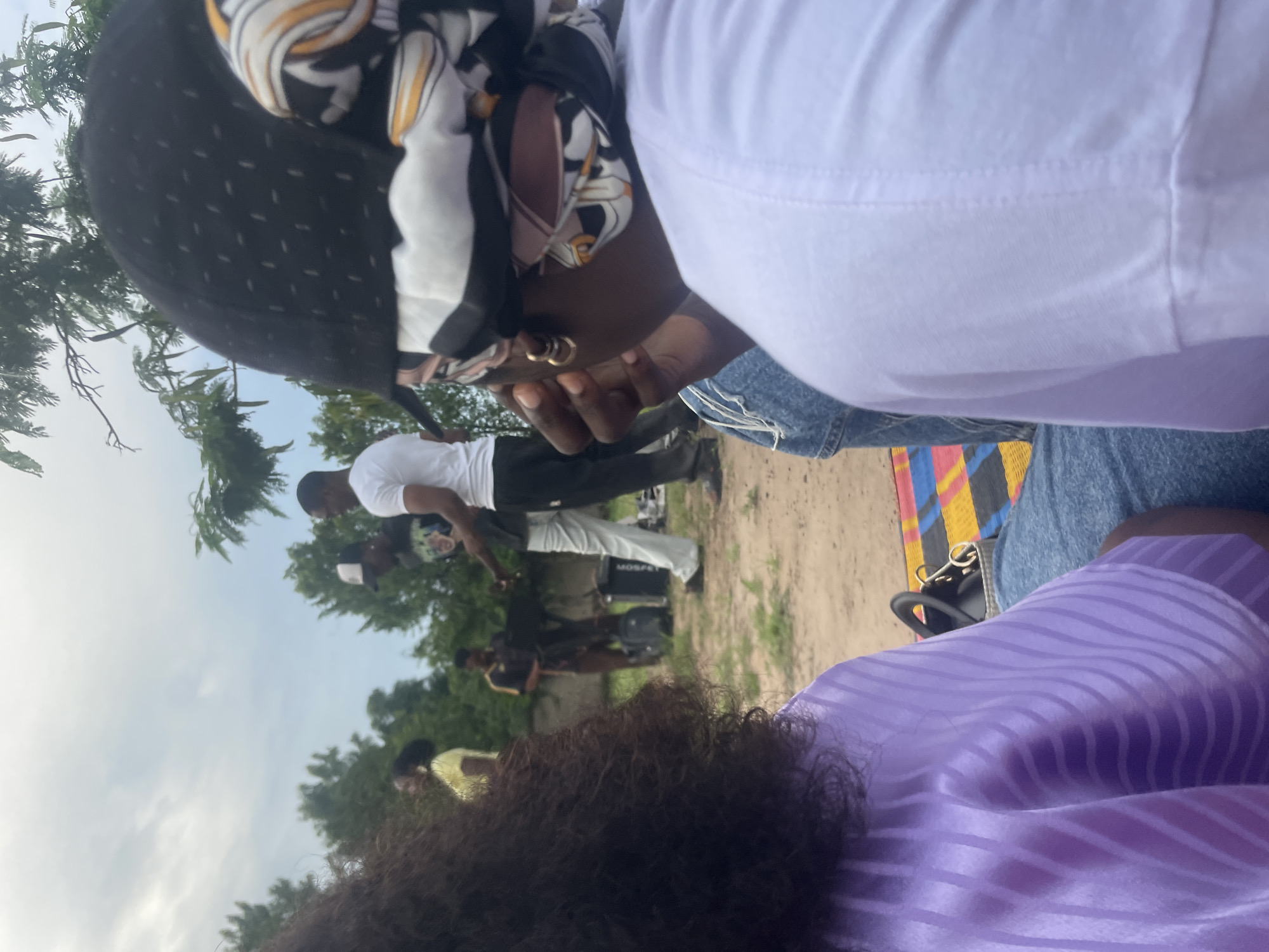 A group of people are gathered outdoors, possibly in a park or an open field, in Lafia, Nigeria. In the foreground, two individuals are seated, with the camera focused on the back of their heads and shoulders. One person on the left has long, dark, curly hair and is wearing a purple, striped garment. The person on the right is wearing a white t-shirt and a headscarf with a black and white patterned design. They appear to be looking towards a group of people in the middle ground.
In the middle ground, three individuals are standing. One man wears a black t-shirt with a graphic print and a white baseball cap. Another man, in a white t-shirt and light-colored pants, appears to be speaking to him. A third person, dressed in dark clothing, is partially visible behind them. They seem to be engaged in conversation or some activity.
The environment is an outdoor setting with dry, sandy ground and scattered green vegetation and trees in the background. The sky is overcast. There are some bags and equipment on the ground near the people in the middle ground, suggesting a gathering or an event. A colorful mat is visible under the two foreground individuals. The overall scene conveys a casual social gathering.
