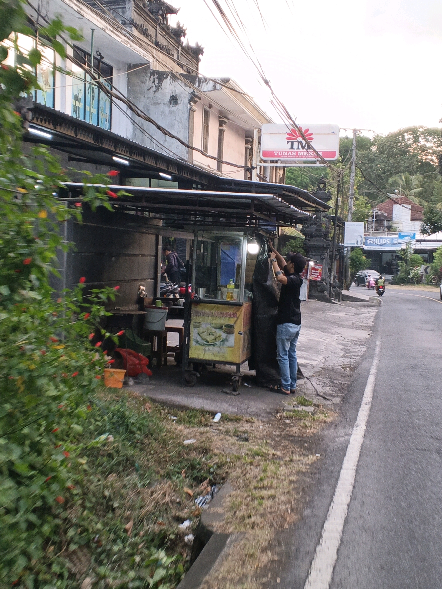 In Manggis, Indonesia, a roadside food stall operates on the right side of a paved road. A man wearing a black t-shirt and blue jeans is in front of the stall, facing away from the camera, and appears to be tying a large black plastic bag to a metal frame. Beside him, a food cart is visible, with a glass display and yellow signage. A second person, partially obscured and wearing a dark jacket, can be seen inside the stall area, behind the food cart. The stall is under a corrugated metal roof supported by metal poles.
The environment includes a building with a weathered facade and traditional architectural elements on the left. Lush green foliage with small red flowers is in the foreground on the left. The road extends into the distance on the right, where two motorcycles are visible. A sign with the logo "TM Tunas Mekar" is mounted on a pole near the building. The ground around the stall is a mix of dirt, grass, and some scattered debris. Overhead, a tangle of power lines crisscrosses the scene.