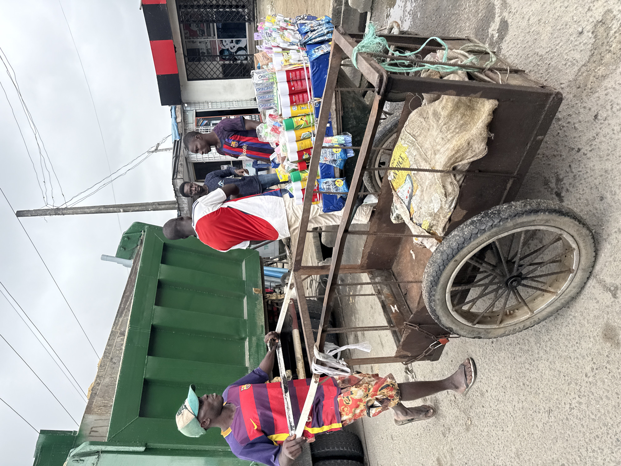 In Ado, Nigeria, an outdoor scene unfolds with a large, green dump truck partially visible on the left. A man wearing a purple and red striped shirt, a green cap, and patterned shorts is pushing a two-wheeled cart. The cart is loaded with various items, including spray cans, packaged goods, and what appears to be bags of rice covered with burlap. Three other men are visible in the background. One man in a red and white shirt is facing away from the camera, while two others stand near a building with red and black accents. The environment appears to be a street or market area, with overhead wires and a cloudy sky.