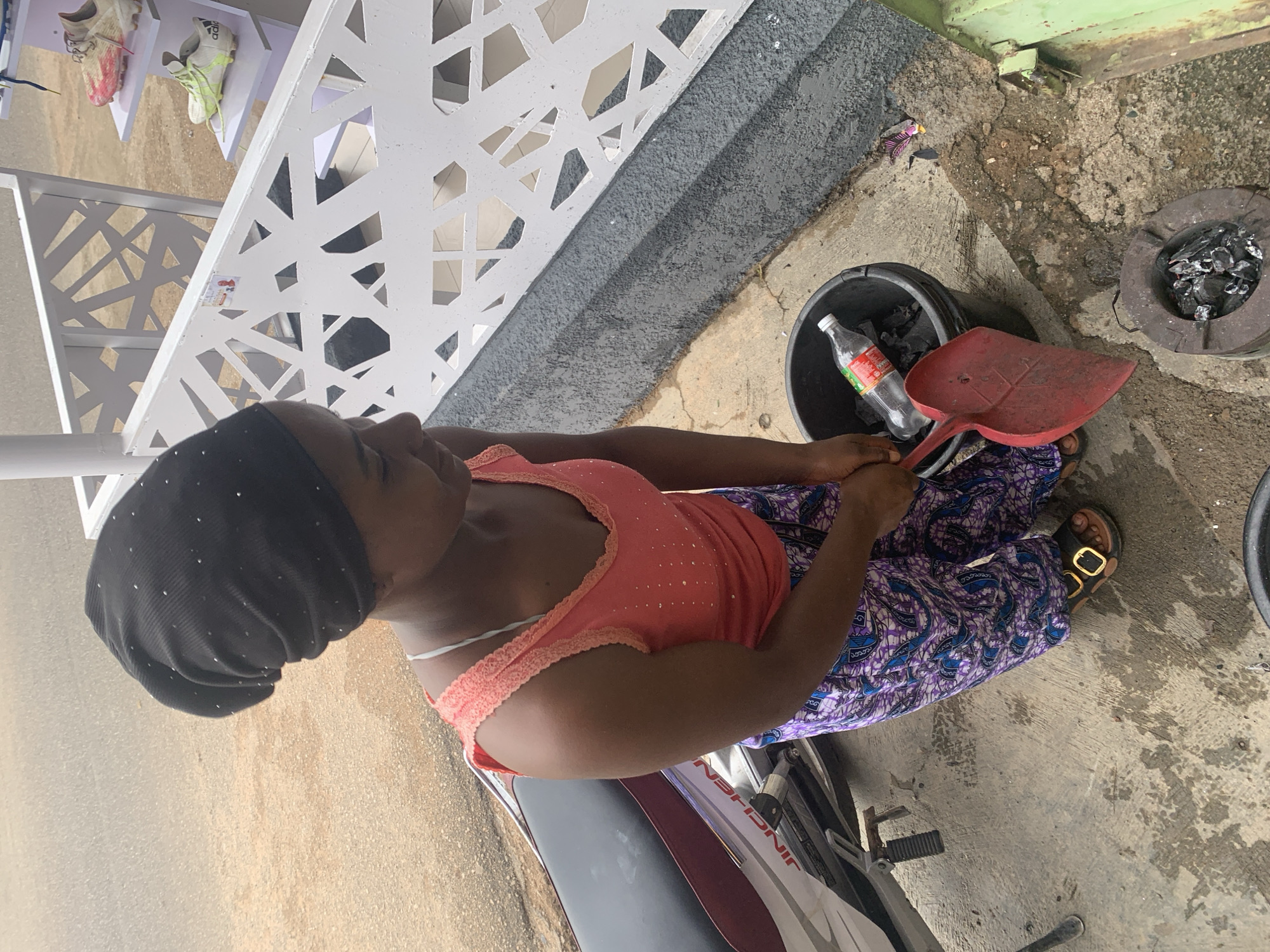 A Black woman wearing a black beanie and a coral-colored tank top with beaded embellishments and a purple patterned skirt is standing next to a motorcycle and a charcoal stove. She holds a red dustpan in her right hand and appears to be transferring charcoal from a black bucket to the stove. A clear plastic bottle with a red label is visible in the bucket. The ground is concrete and appears wet in places. In the background, a white decorative railing with an abstract pattern separates the walkway from a display of shoes on white shelves. The scene suggests an outdoor setting in a commercial area of Jos, Nigeria.