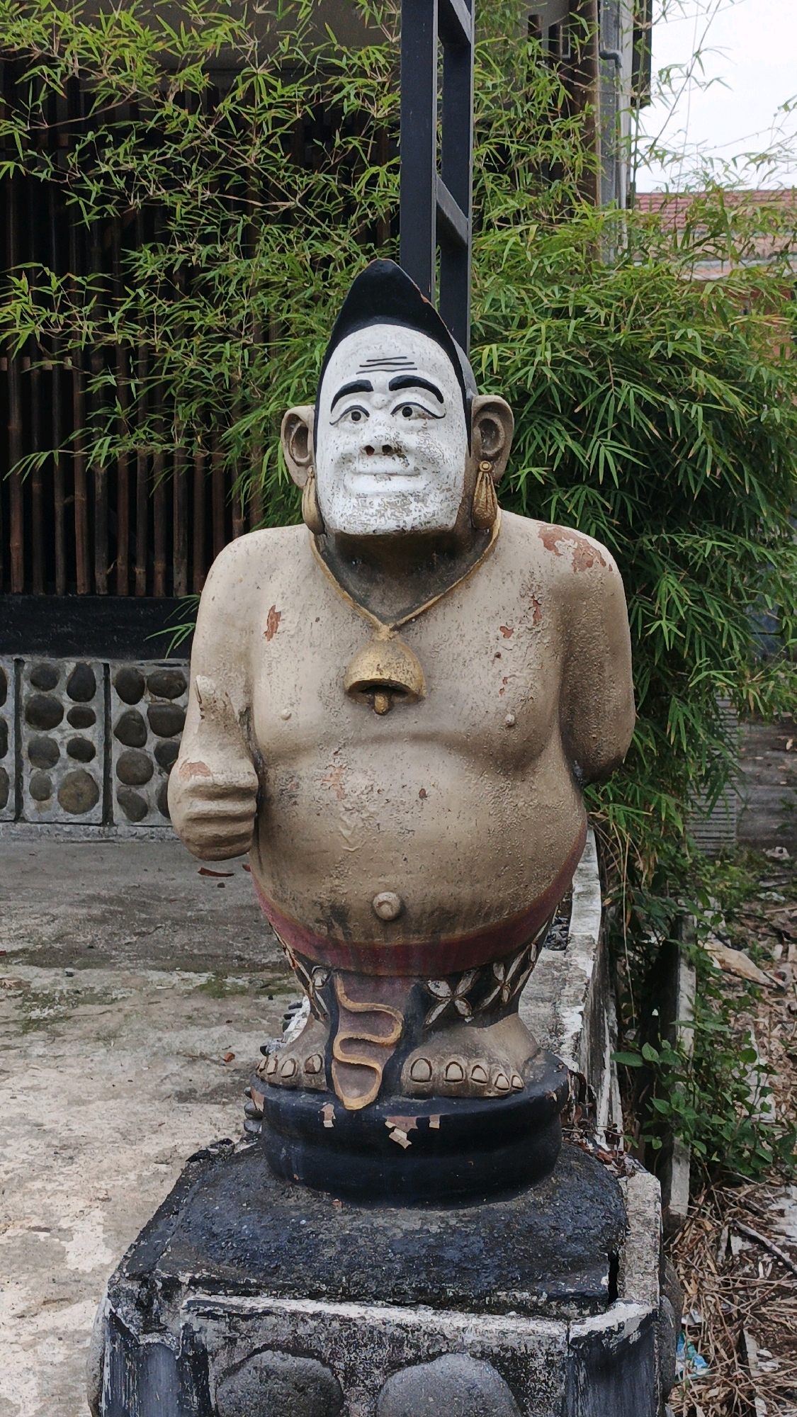 This is a vertical, medium shot of a weathered statue of a stout, humanoid figure with an exaggerated, grinning face. The statue is positioned outdoors, on a concrete pedestal. The figure has a white face with large, round eyes and prominent eyebrows. It wears what appears to be a black, pointed headpiece. Large, golden earrings hang from its ears, and a golden bell on a cord rests around its neck. The body is beige and shows signs of peeling paint and texture. One arm is bent at the elbow, with the hand raised in a thumb-up gesture. The lower body, which resembles legs, is adorned with intricate, dark patterns and the figure stands on a black, circular base.
Behind the statue, dense bamboo foliage creates a green backdrop. To the left of the statue, a wall constructed with rough-hewn stones is visible. The ground in front of the statue is a patchy, light-colored concrete. The overall setting suggests an outdoor space, possibly a garden or courtyard, in a tropical or subtropical environment, given the presence of bamboo and lush greenery. The condition of the statue indicates it has been exposed to the elements for a considerable time. The image was taken in the city of Sukaresik, Indonesia.