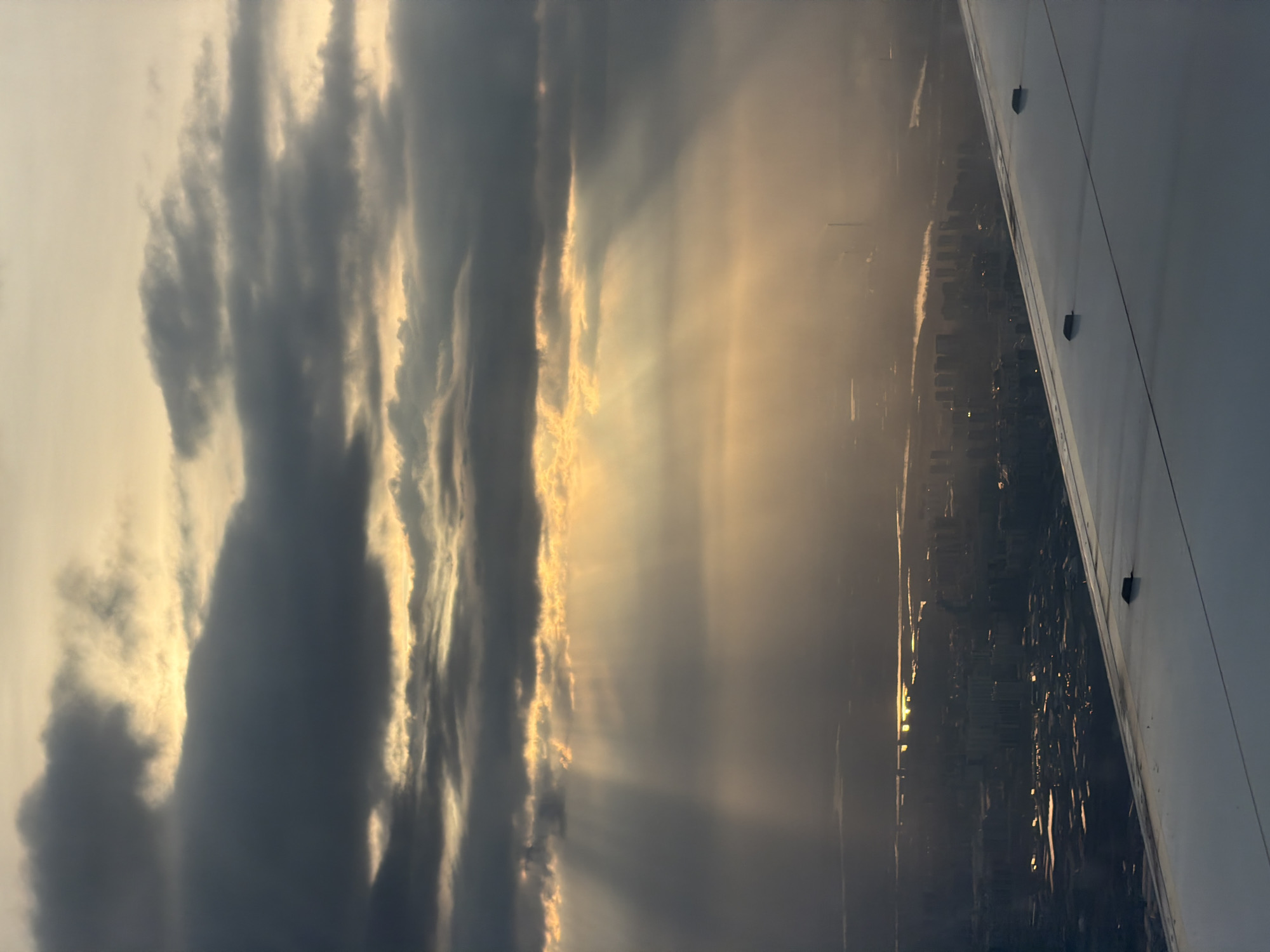 The image shows a view from an airplane window. The wing of the aircraft is visible on the right side. In the background, a city skyline is partially obscured by clouds and light rays. The lighting suggests either sunrise or sunset. Based on the urban layout and atmospheric conditions, the city is likely Krasnoyarsk, Russia. No people are visible. The scene appears to be a combination of natural atmospheric phenomena and urban development viewed from an elevated perspective.
