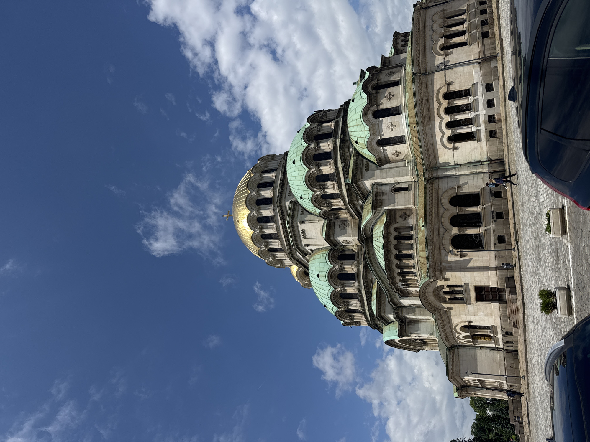 The image captures a vibrant daytime scene dominated by the Alexander Nevsky Cathedral in Sofia, Bulgaria, under a clear blue sky with scattered white cumulus clouds. The grand Eastern Orthodox cathedral, with its light-colored stone facade, features a prominent golden central dome and numerous smaller, intricately designed domes and roofs that have oxidized to a distinctive verdigris green. The cathedral's architecture includes multiple tiers, arched windows, and detailed stonework, with strong sunlight casting clear shadows that highlight its relief.
On the paved ground in front of the cathedral, two small figures of people are visible walking, suggesting pedestrian activity. A dark-colored car is partially visible on the far right edge of the frame, indicating proximity to a street or parking area. Small green shrubs or potted plants are also visible along the paved pathway near the building. The overall impression is one of a bright, sunny day at a historic and impressive architectural site. No visible text is present in the image.