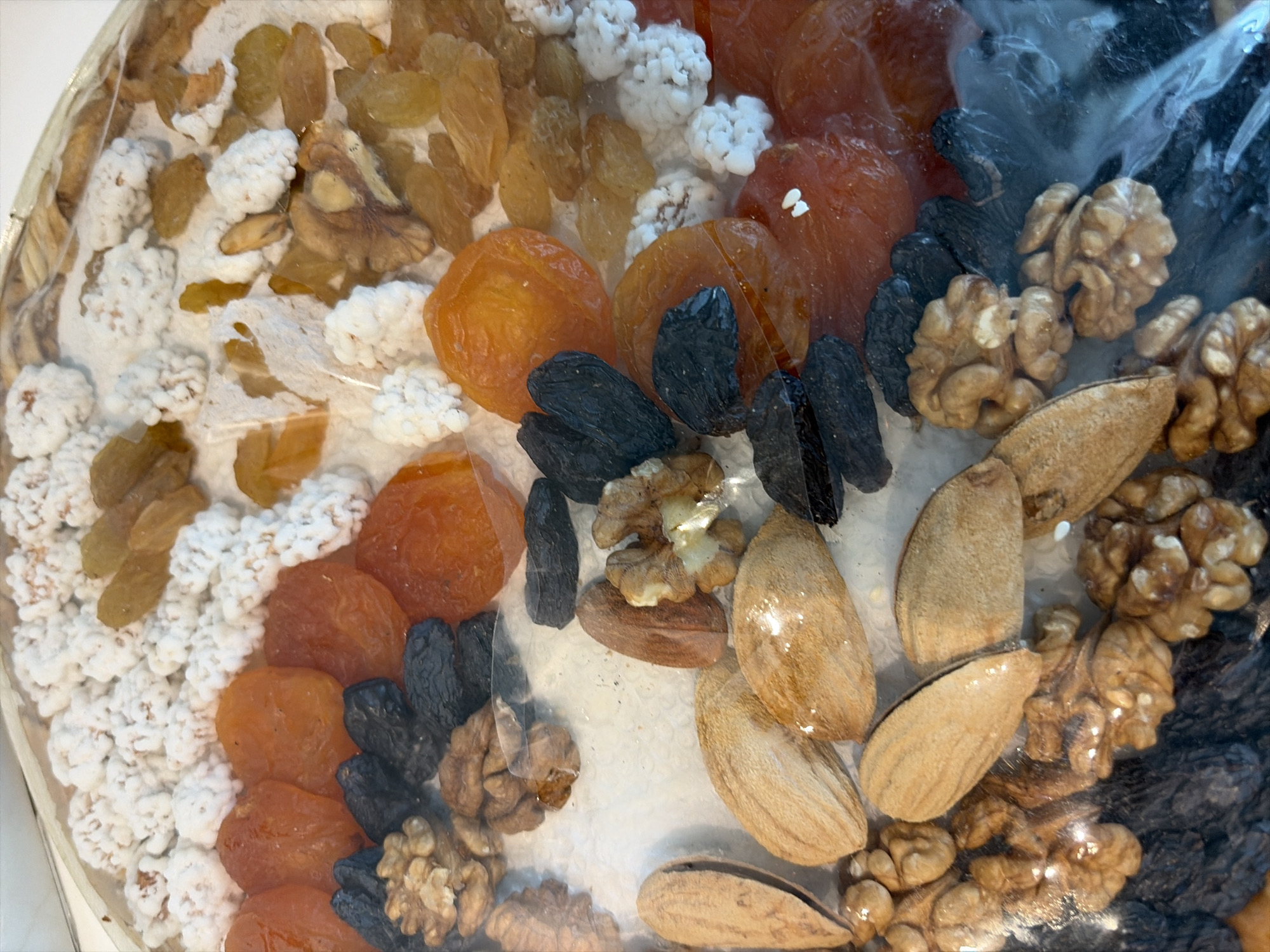 The image is a close-up, overhead view of a round assortment of dried fruits and nuts contained within clear plastic packaging. The contents include golden raisins, dried apricots, black raisins, walnuts, and almonds, as well as small, irregular white clusters that appear to be sugar-coated nuts or candies. The items are densely packed and arranged in a somewhat random, decorative pattern. The lighting creates some reflections on the plastic. There is no indication of a specific location within Moscow, Russia, or any people or actions.