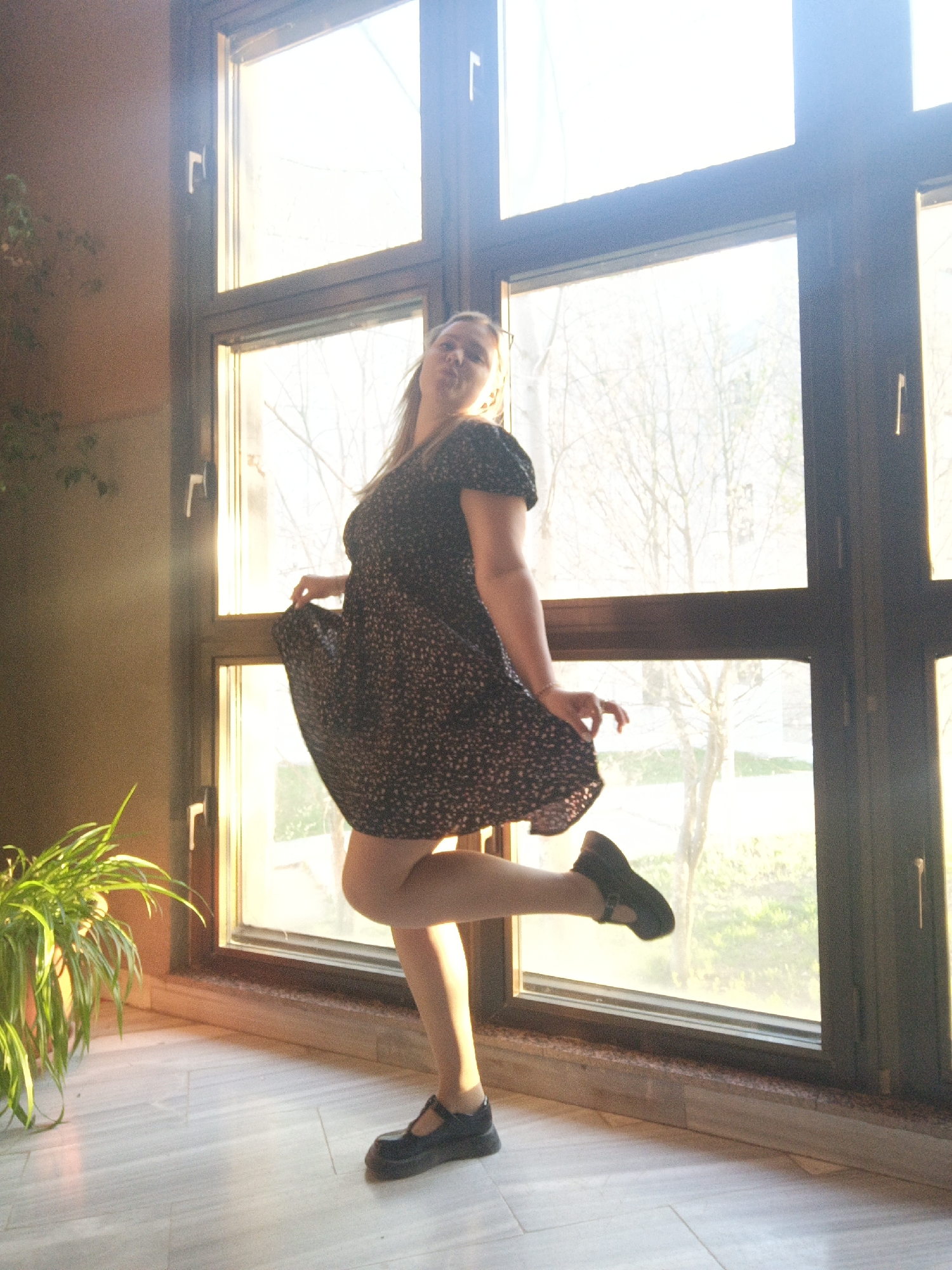 A woman in a dark, floral-patterned dress and black Mary Jane shoes is standing near a large window. She is lifting the skirt of her dress with her left hand, and her right leg is slightly raised. A potted plant is visible on the left side of the frame. The window looks out onto a scene with bare trees and some green vegetation, suggesting an outdoor setting, with possible light coming from the city of Markovskii, Russia.