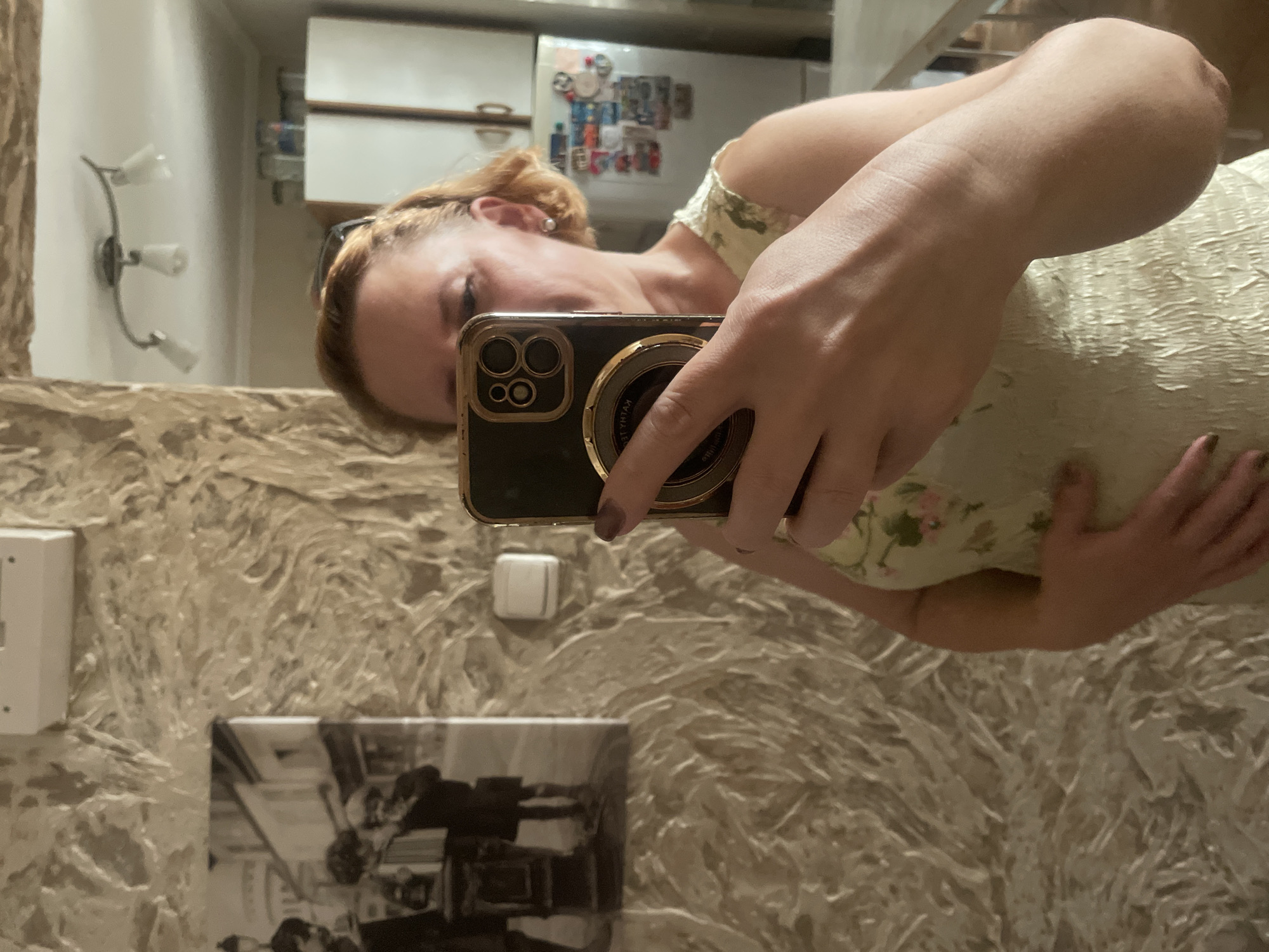 The image is a mirror selfie taken by a person from a slightly elevated angle, with the camera held in their right hand. The subject is a woman with blonde hair pulled back, wearing a light-colored dress with a floral pattern. She is leaning back, and her face is partially visible as she looks towards the camera. Her right arm is extended, holding a smartphone with a gold-accented camera module. The reflection shows a bathroom or kitchen environment, with cabinets and a light fixture visible in the background. The wall has a textured, stone-like finish. A framed picture is visible on the wall below the light fixture. The lighting appears to be artificial and somewhat dim. The overall impression is a casual, personal snapshot.