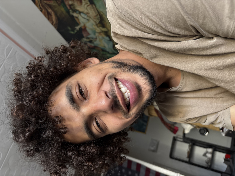 A close-up portrait features a young man with dark, tightly curled hair and a dark beard and mustache, smiling widely at the camera. His white teeth are visible, and his eyes are crinkled at the corners, conveying a joyful expression. He is wearing a plain, light brown or tan short-sleeved t-shirt. The photograph appears to be a selfie taken from a slightly high angle.
The background, though somewhat blurred, provides glimpses of an indoor setting. Behind the man's head, an ornate framed artwork or tapestry, possibly depicting classical figures, is visible on the wall. To the lower left of the frame, a distinct American flag is partially visible, either draped or folded as a decorative element. Other indistinct elements of a room, such as furniture, can be seen further in the background to the right. The lighting suggests it is daytime. The presence of an American flag in Tangier, Morocco, is a notable detail, potentially indicating cultural ties, a themed space, or personal decor. No discernible text is visible in the image.