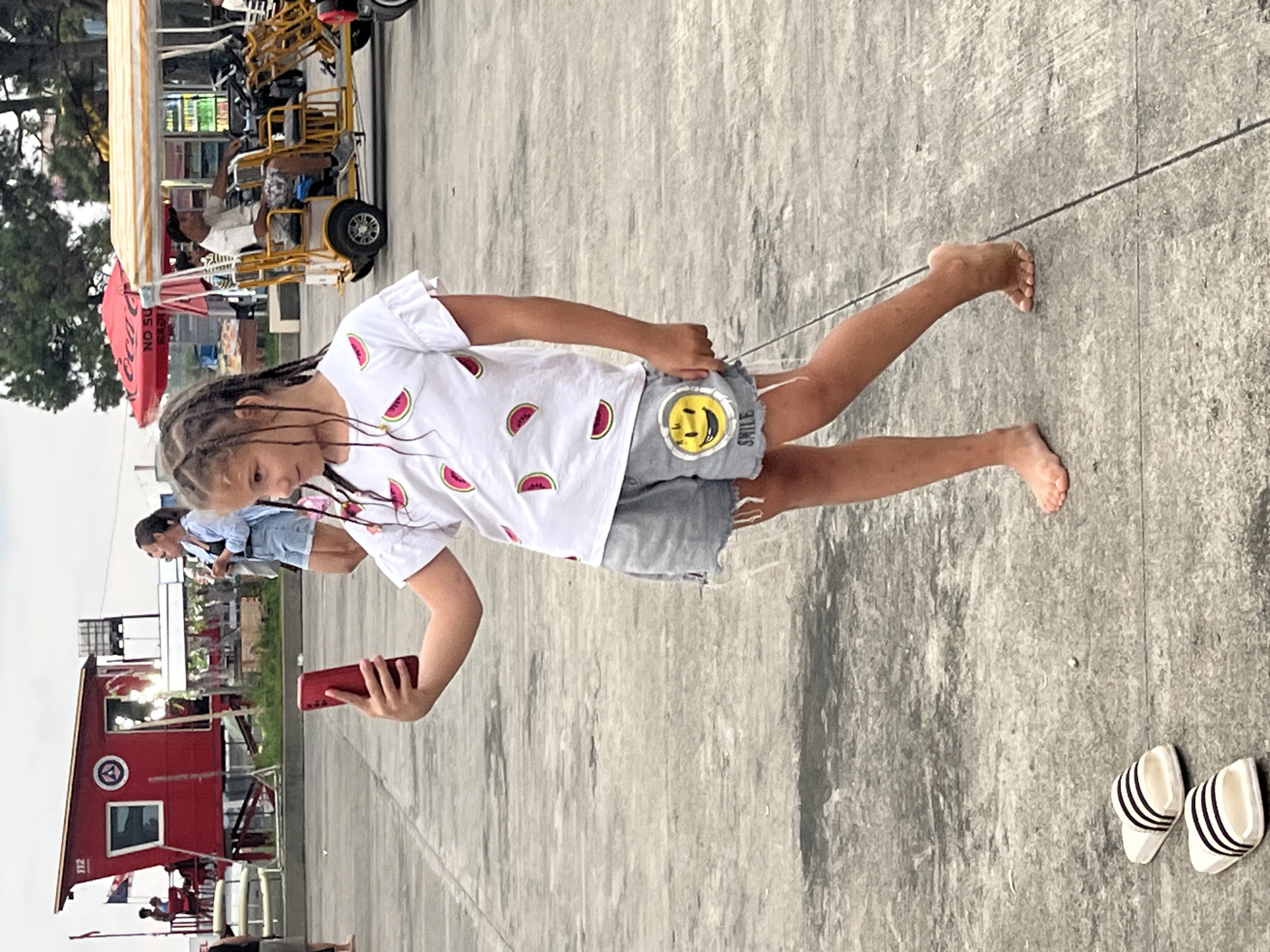 A young girl with braided hair is standing on a paved area, holding a red smartphone to take a selfie. She is wearing a white t-shirt with watermelon slices printed on it and gray denim shorts with a yellow smiley face patch and the word "SMILE" printed below it. Her left foot is raised, and her right foot is flat on the ground. To her left, on the ground, are a pair of white flip-flops with black stripes. In the background, to the left, is a red lifeguard tower with the number "112" on it. Further in the background, there are people, a yellow tourist cart, and red umbrellas. The scene appears to be a public area, possibly near a beach, consistent with the resort town of Ureki, Georgia, known for its magnetic sand beaches and recreational facilities.