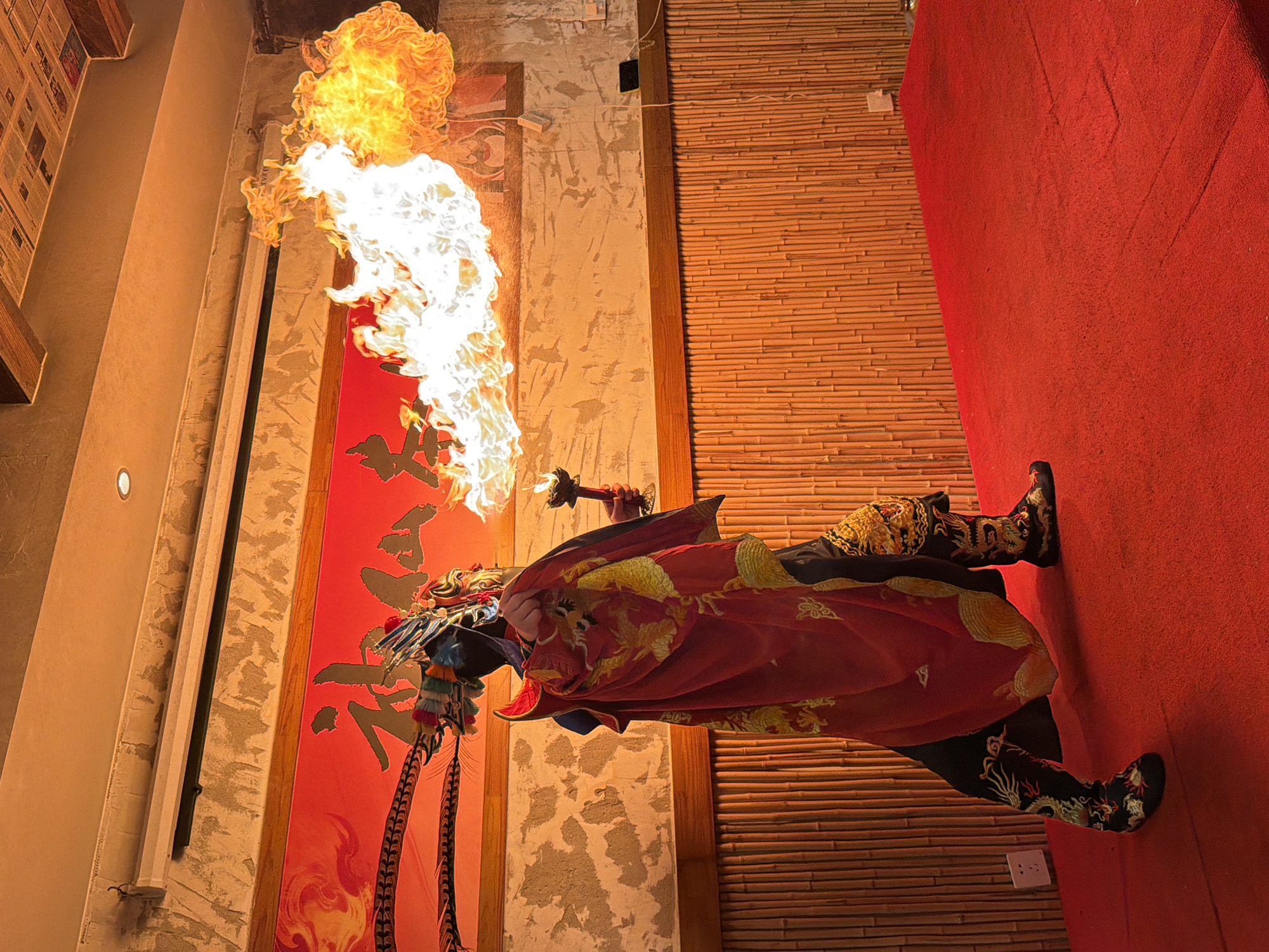 An individual in a theatrical costume, likely a performer of traditional Chinese opera or a similar act, is on a red stage in what appears to be a performance space. The costume is elaborate, featuring a red cape with gold dragon embroidery, a headdress with colorful feathers, and intricately decorated boots. The performer is holding a torch in their right hand, from which a large plume of fire is erupting outwards and upwards, illuminating the scene. Behind the performer is a red banner with stylized Chinese characters and decorative elements that resemble feathers. The backdrop consists of a wall covered in vertical bamboo stalks. The floor is a vibrant red carpet. The overall atmosphere is one of dramatic performance, possibly in a cultural venue or restaurant. The location is identified as Yuzhong District, China.