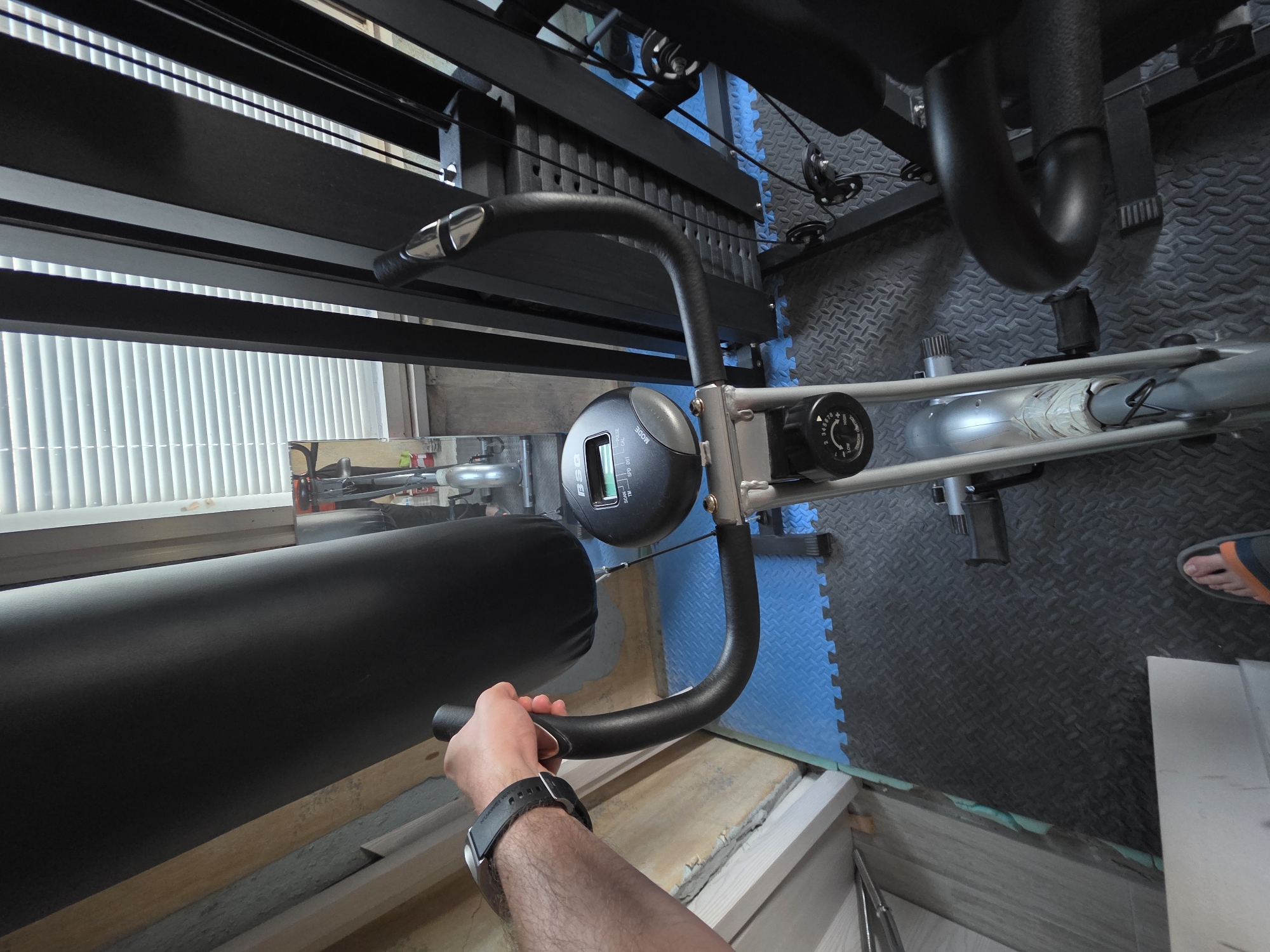 The image depicts a person holding the handle of a piece of exercise equipment, possibly an ab machine or similar fitness apparatus. The equipment has a padded black cushion, a black metal frame, and a digital display screen on a control panel. The person's forearm, wearing a watch, is visible. The equipment appears to be located in a room with interlocking foam floor tiles, partially blue and partially black. There's also what looks like a stationary bicycle in the background. Light is entering through a window with blinds, suggesting an indoor setting. The location in Yagodovo, Bulgaria cannot be determined from the visual data.
