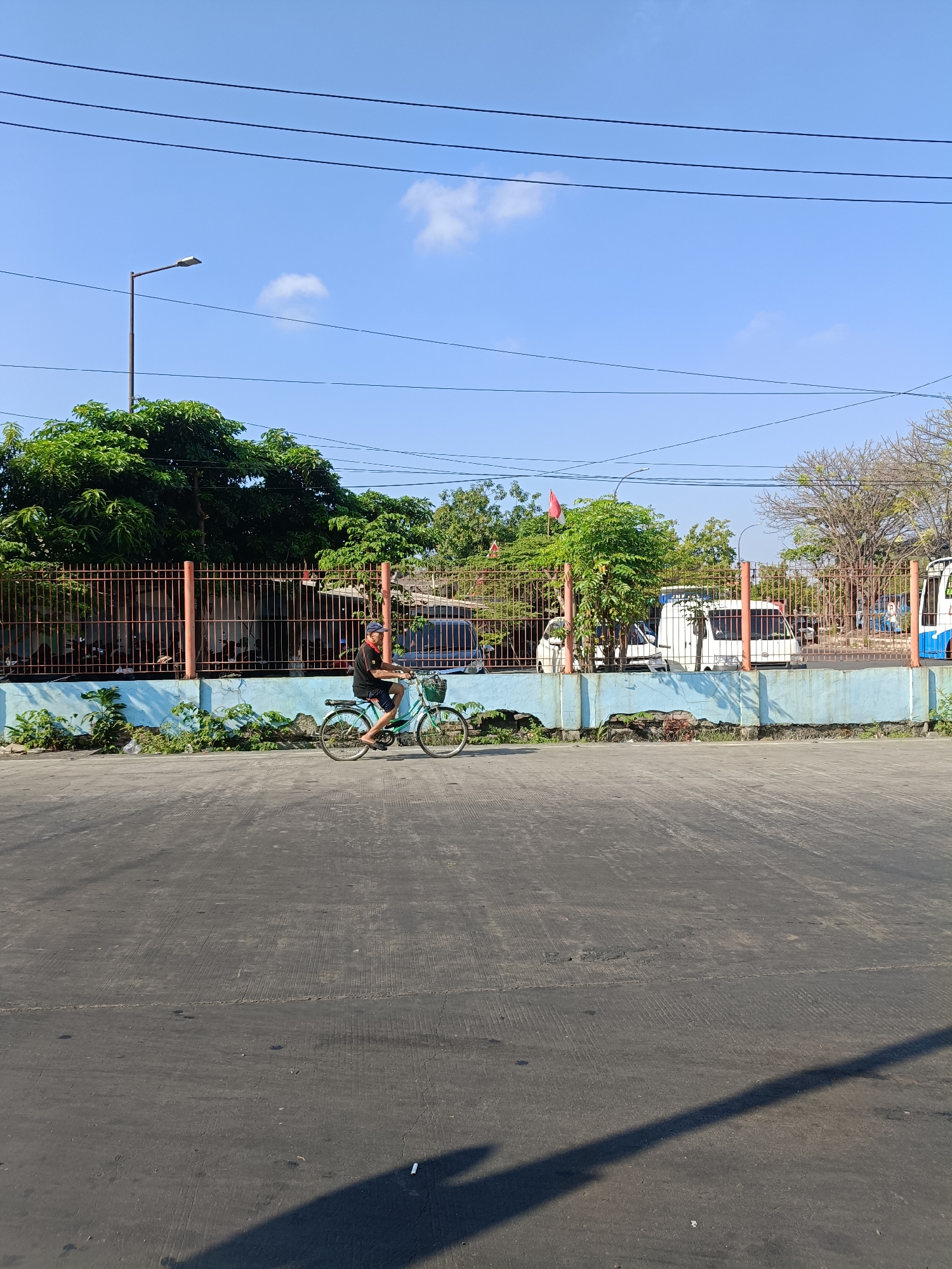 A man rides a bicycle on a paved road in the city of Kedungrejo, Indonesia. He is wearing a black t-shirt, blue shorts, and a blue cap. The bicycle is light blue and has a basket on the front. The man is in motion, and his shadow is cast on the road behind him. The road is asphalt and shows signs of wear, with tire marks and some cracks.
In the background, there is a blue and orange fence, behind which are several vehicles, including white vans and what appears to be a bus. Trees and foliage are visible above and behind the fence, and a street lamp stands on the left side of the image. The sky is clear and blue with a few scattered clouds. The overall scene suggests a bright, sunny day.
