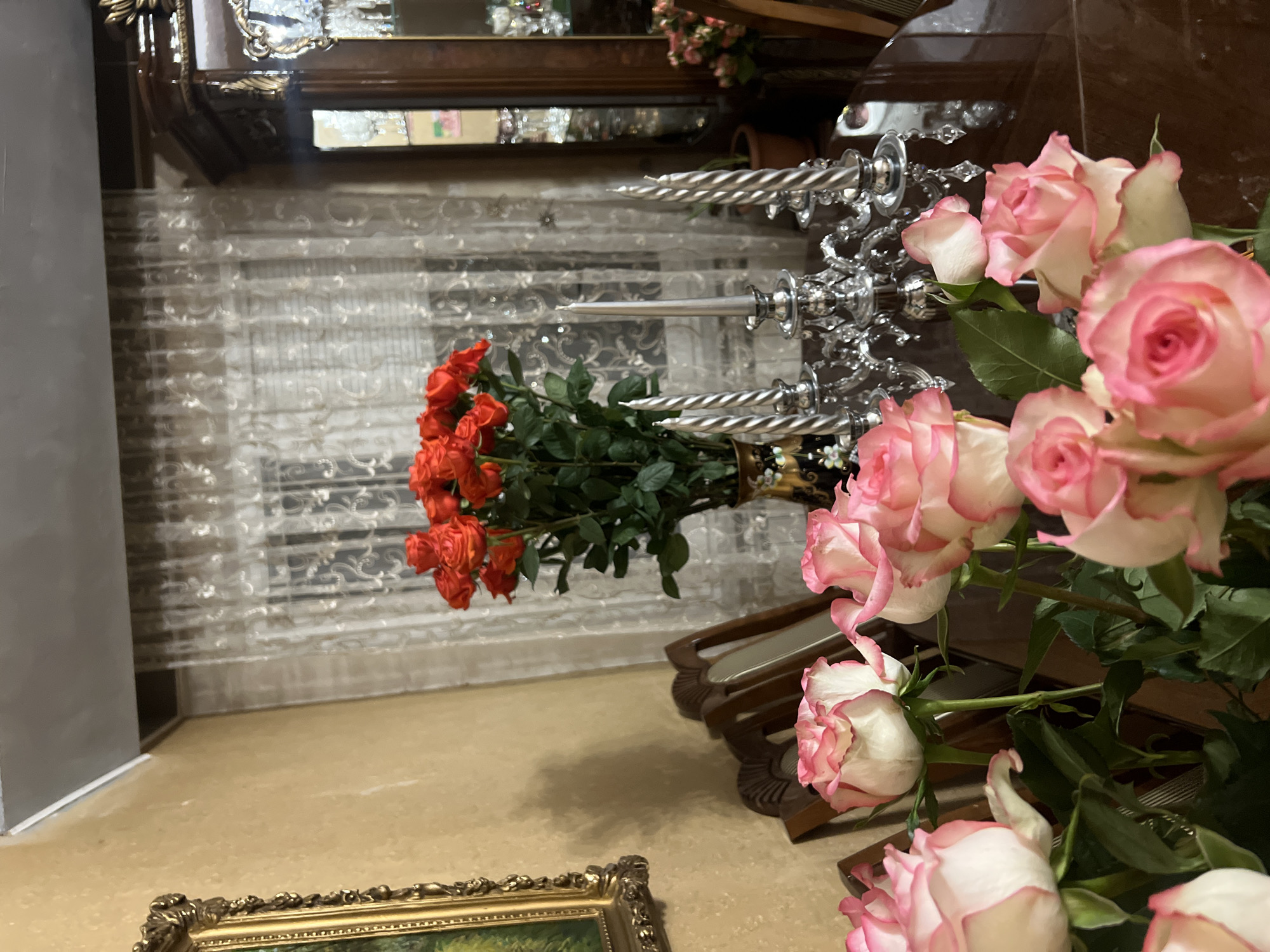 Indoor shot. An ornate silver candelabra stands between a bouquet of red flowers and a bouquet of pink and white roses. A dark ornate vase holds the bouquets. The foreground includes a gold framed painting. A mirror is in the background, reflecting part of the scene. The environment appears to be a home. The location is Yerevan, Armenia.