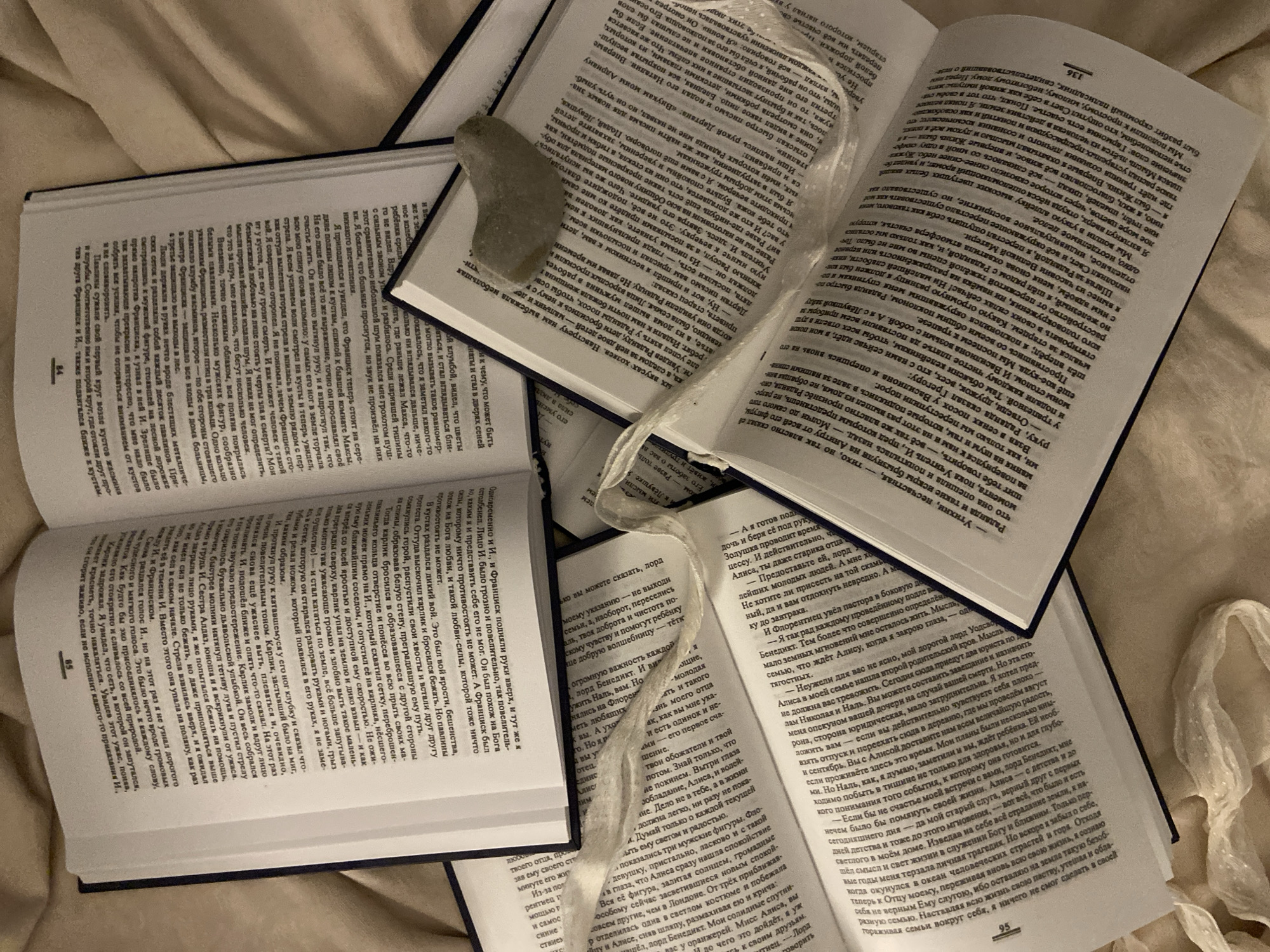 This indoor scene is located in Budva, Montenegro. Multiple hardback books, bound in dark blue with white pages, are lying open and scattered across a light beige, soft textile surface, likely a bed or blanket. The visible pages display dense Cyrillic text, with page numbers such as "85," "84," "136," and "95" discernable. A small, smooth, pale gray stone of irregular shape rests on the upper right page of one book. A length of crumpled, translucent white material, resembling a thin plastic or synthetic ribbon, is draped loosely across several of the open books. No individuals are present in the image. The arrangement suggests a casual reading environment.