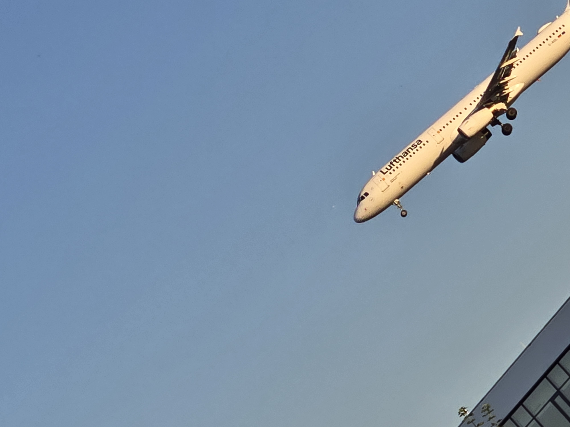 A Lufthansa airplane, identified by the logo on its fuselage, is captured in mid-flight against a clear blue sky. The aircraft appears to be on its approach or departure, as its landing gear is extended. The sun is casting a warm light, suggesting it is either early morning or late afternoon. In the lower right corner of the image, a modern building with reflective windows is partially visible, hinting at an urban or airport setting. The overall scene conveys a sense of travel and transit. The visible text includes "Lufthansa" on the side of the plane, and a registration code that appears to be "D-AIS..." though it is partially obscured. The image is taken from a low-angle perspective, looking up at the airplane. The weather is clear with no clouds visible.