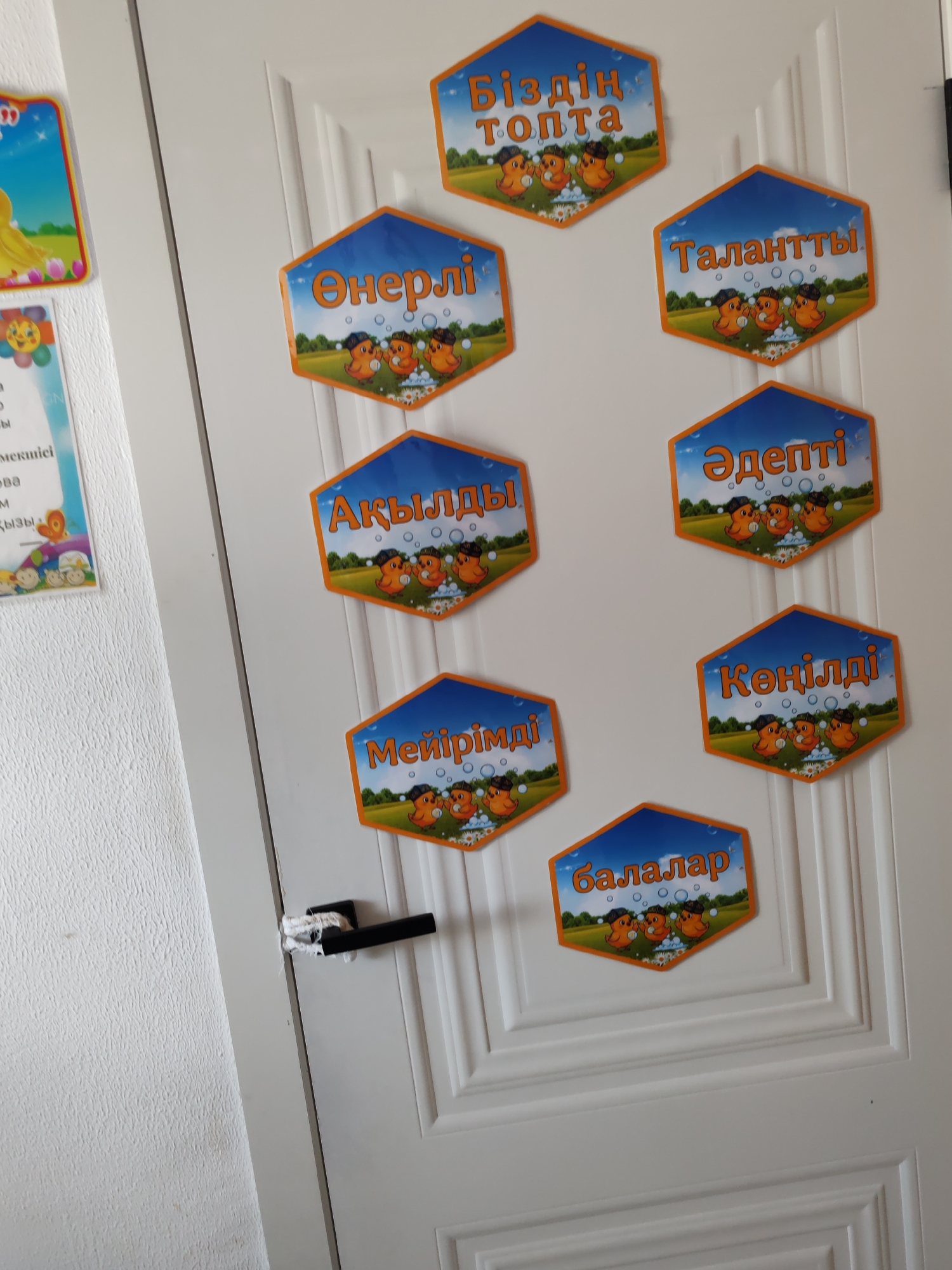 The image shows a white door with several hexagonal-shaped posters attached to it. The posters have an orange border and a blue sky background with clouds and green grass. Each poster features an illustration of three yellow chicks wearing black hats and standing in bubbles. The text on the posters is in Kazakh.
The posters read:
- "Біздің топта" (Our group)
- "Өнерлі" (Talented/Creative)
- "Талантты" (Talented)
- "Ақылды" (Smart/Wise)
- "Әдепті" (Polite/Well-mannered)
- "Мейірімді" (Kind)
- "Көңілді" (Cheerful/Happy)
- "Балалар" (Children)
To the left of the door, on the textured wall, are two other posters. One poster features a yellow duckling in a bright, colorful scene. The other is partially visible and appears to have text and images related to children.
A black door handle is visible on the door, secured with white string or thread. The overall scene appears to be in a classroom or children's area. The presence of the Kazakh language and the specific educational themes on the posters suggest a location in Kazakhstan, likely a school or kindergarten. Given the detail, this specific arrangement of posters on a door is unlikely to be precisely pinpointed to a specific street in Aktzhar, Kazakhstan without further context or geographically unique identifiers within the image itself.