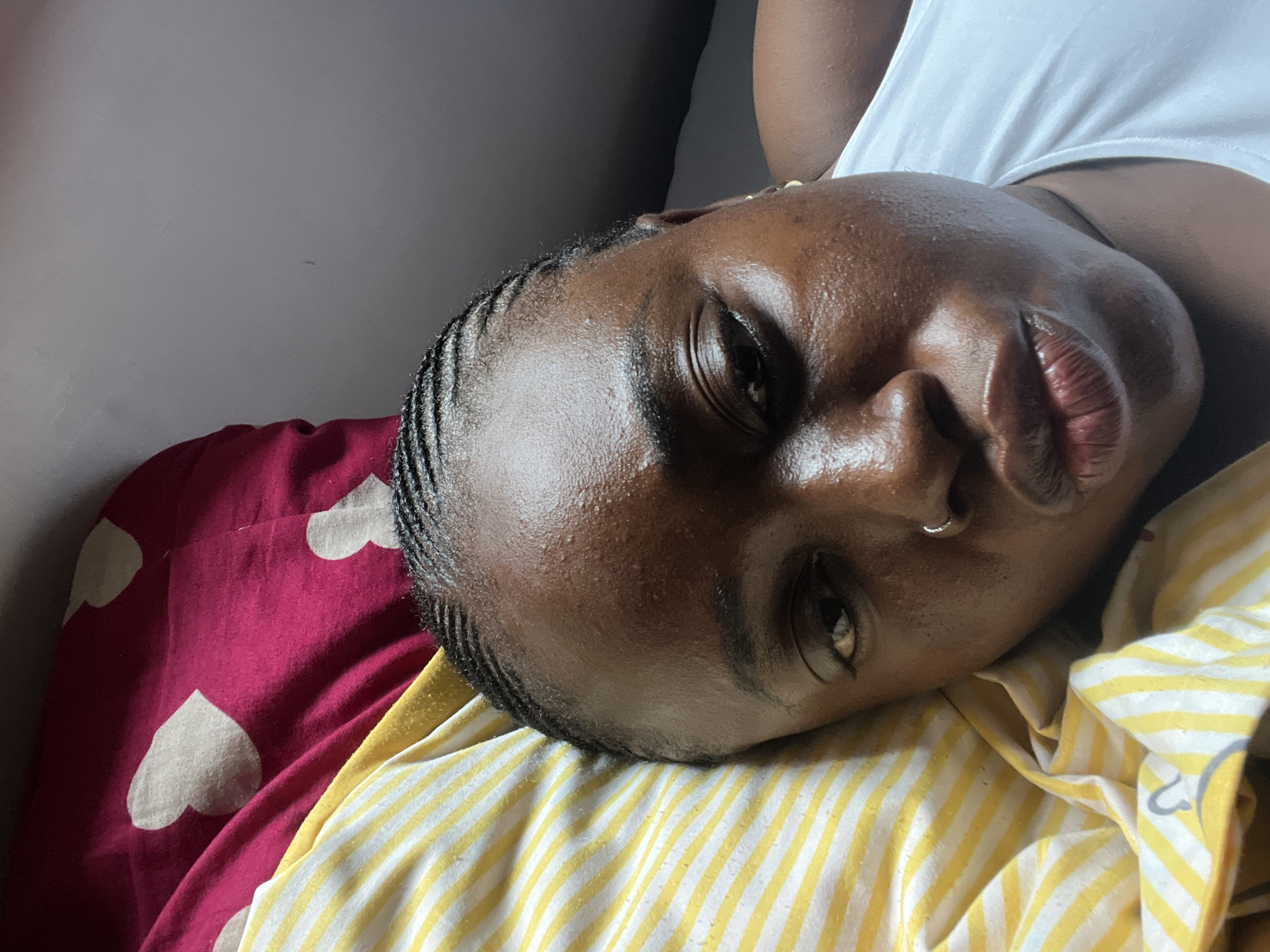 A close-up, overhead shot captures a dark-skinned woman lying on her side in a bed. She has her eyes closed and her lips pursed, as if posing for a kiss. Her hair is styled in cornrows, and she is wearing a white tank top. The background is a blurred mix of yellow and white striped fabric and a pink fabric with white hearts. The setting appears to be a bedroom in Abuja, Nigeria. The lighting is soft and diffused, suggesting natural light from a window. The overall mood is intimate and relaxed.