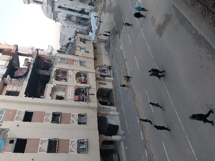A partially destroyed building stands adjacent to a mosque with a white dome. The building's structure is severely damaged, revealing interior spaces. An asphalt road runs alongside the building, with multiple people walking on it, some moving in groups and others individually. The scene suggests urban devastation in the aftermath of conflict.
