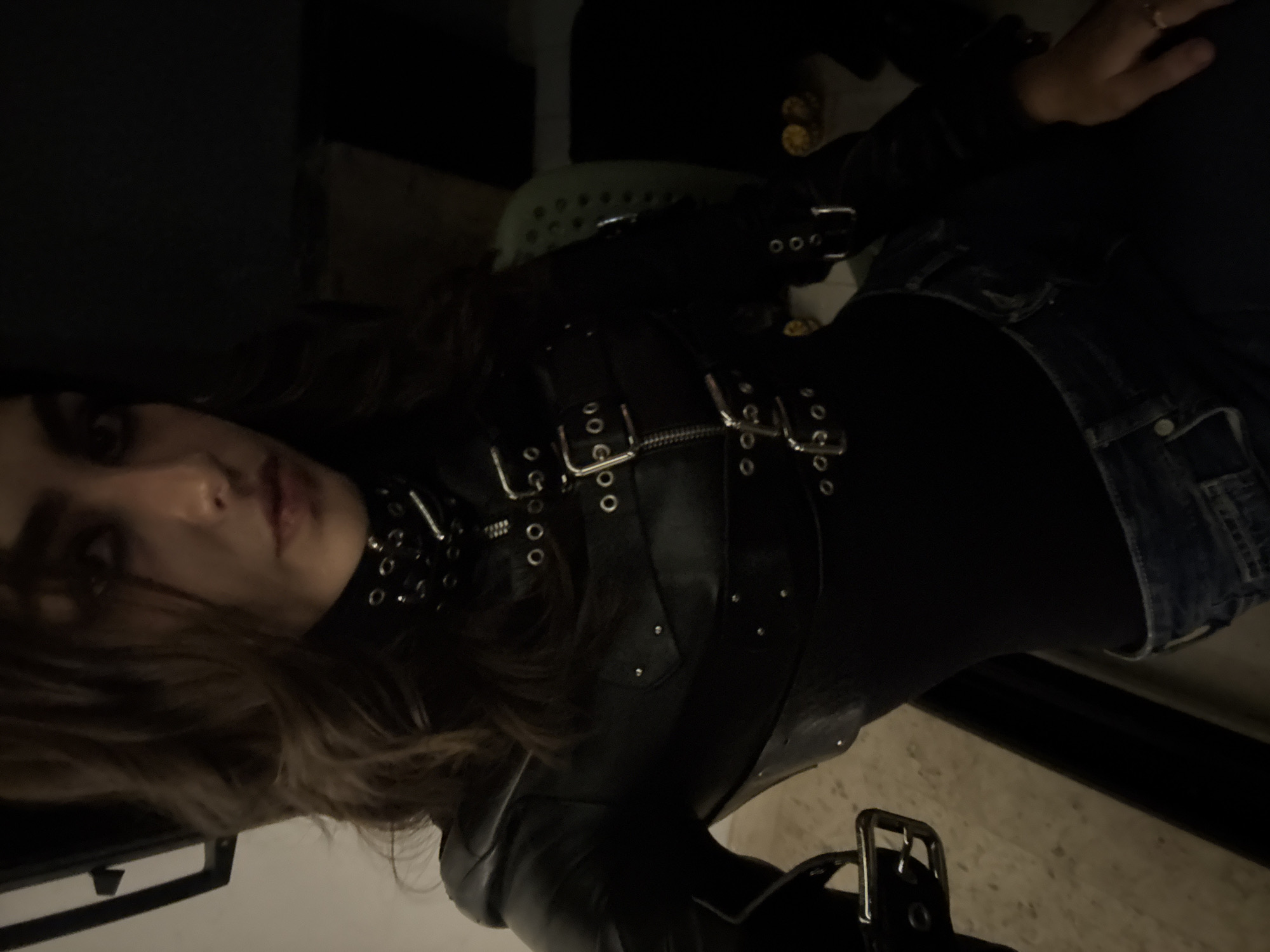 A young woman is shown in a selfie-style shot. She is wearing a black leather harness-like garment with metal buckles and rivets. She has on a black turtleneck shirt and denim jeans. The location is not clear from the image and cannot be confidently identified as Mexico City. The lighting is low, suggesting an indoor setting.
