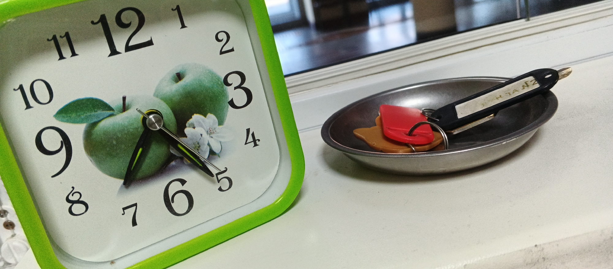 The image shows a close-up of a green-framed analog clock with a depiction of green apples. The clock face has black numerals and hands, with the time appearing to be approximately 9:47. To the right of the clock is a small, metallic, shallow dish containing two keys with keychains. One keychain is red and heart-shaped, and the other is tan and irregularly shaped. The keys and keychains are resting on the dish, which is positioned on a white surface, possibly a windowsill. The background is blurred, showing reflections on a window. The image does not provide any information about the city of Smila, Ukraine, or any specific location within it.