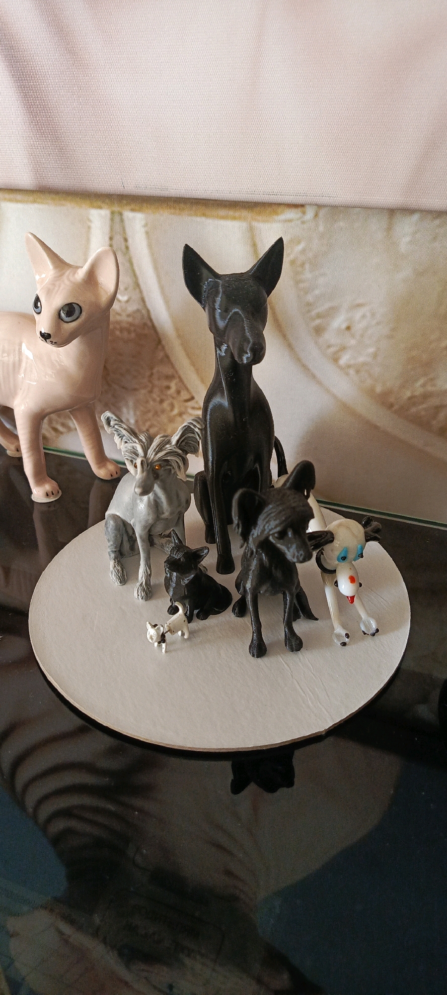 A collection of small figurines of dogs and a cat are arranged on a circular white surface. The figurines include a pink cat with blue eyes, a large black dog, a grey dog with shaggy hair, a small black dog, a tiny white and black dog, a black dog with curly hair, and a white dog with blue eyes and a red tongue. The white circular surface is placed on a dark, reflective surface, possibly a table. In the background, a textured, light-colored wall or fabric is visible, with a blurry image or pattern behind it. The overall lighting suggests an indoor environment. There is no identifiable information in the image to suggest a location in the city of Bolshoy Kamen, Russia.