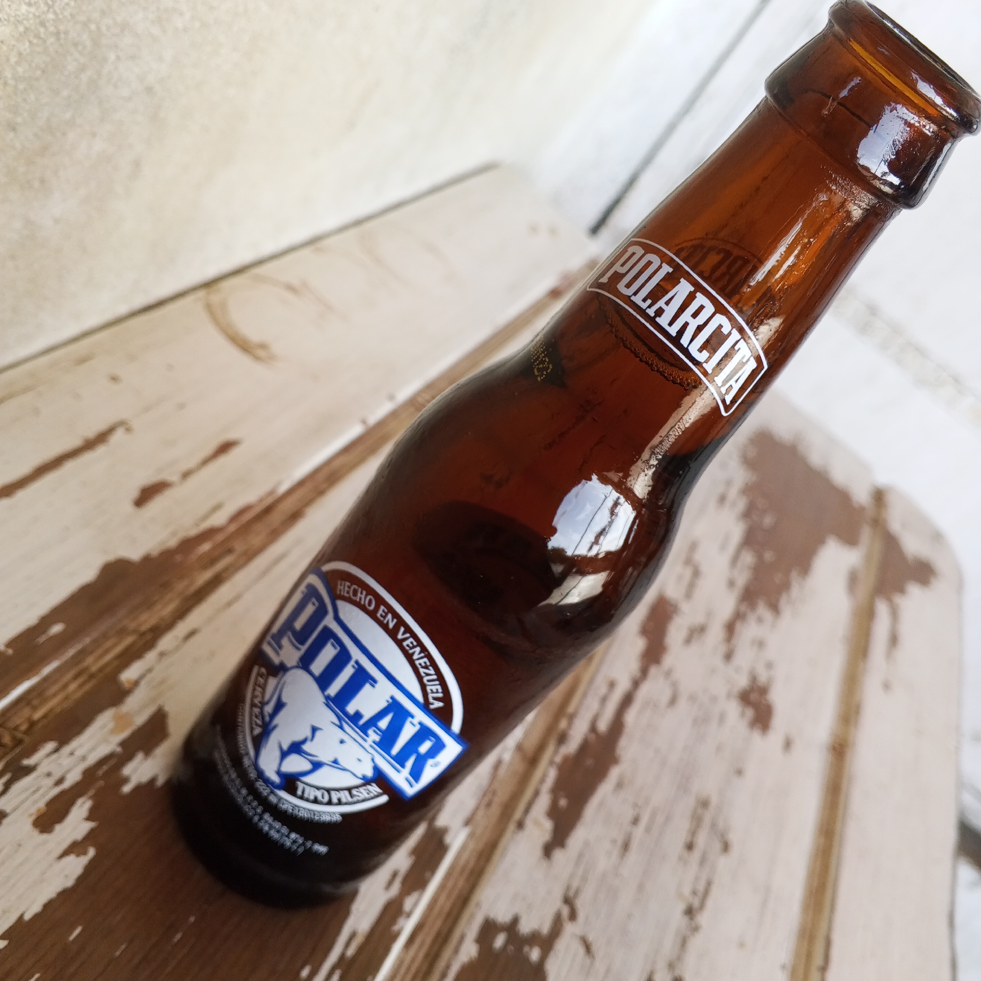 A brown glass beer bottle, branded "POLARCITA" on its neck, rests diagonally on a rustic, distressed wooden surface. The surface appears to be light-colored wood, possibly painted white, with significant wear and tear revealing darker wood grain underneath. In the blurred background, another light-colored surface is visible, suggesting an indoor or outdoor setting.
The bottle features a main label with a blue and white design centered around a polar bear logo. Above the polar bear, the word "POLAR" is prominently displayed. Curved above this, smaller text reads "HECHO EN VENEZUELA" (Made in Venezuela). Below the polar bear, "TIPO PILSEN" is visible, indicating the beer type. Reflections on the glossy bottle surface suggest bright, natural lighting, likely daytime. No people are present, and the bottle is simply placed on the worn wooden surface, conveying a casual atmosphere. The contents of the bottle are not visible due to the dark glass, but it appears full.