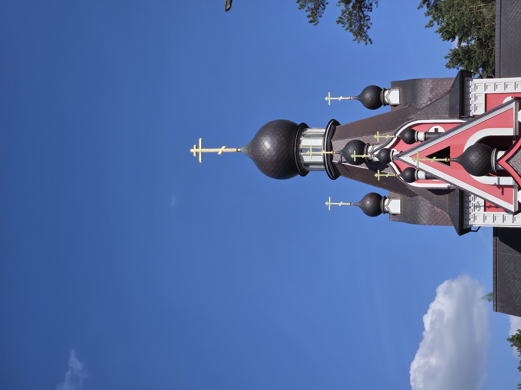 The image shows a Russian Orthodox church with multiple onion domes and crosses against a clear blue sky with a few clouds and treetops. The church is painted in red and white with black domes, and the crosses are gold. The location of the church is Unknown, Unknown.