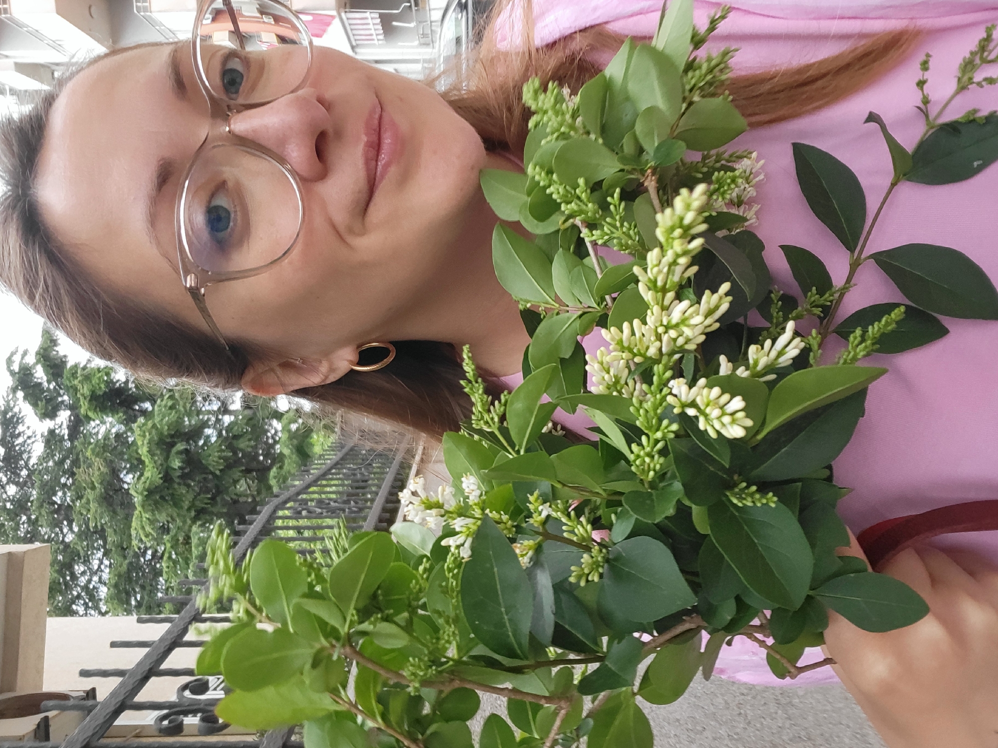A woman with light brown hair and fair skin, wearing round glasses, a light pink shirt, and gold hoop earrings, holds a leafy branch with small white flowers. The background includes an iron fence, green trees, and a building. The location is Budva, Montenegro.