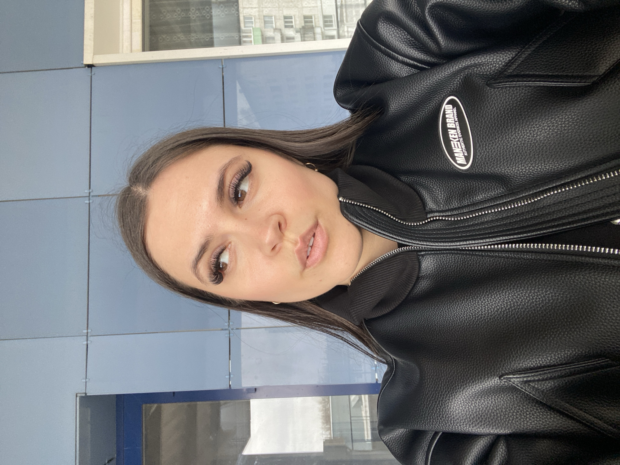 A young woman with long dark hair and artificial eyelashes is posing for a selfie. She is wearing a black leather jacket with a visible logo patch that reads "MANEKEN BRAND AUTHENTIC OVERSIZE APPAREL." The background appears to be a wall made of light blue rectangular tiles with a window. The picture was taken in Chaikovsky, Russia.
