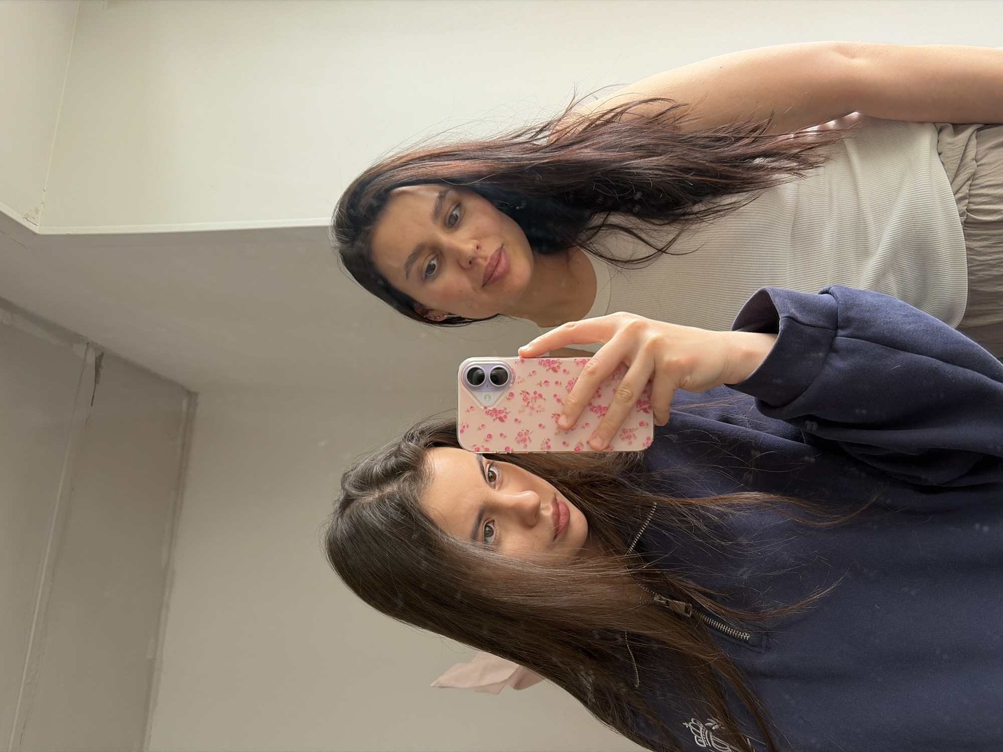 This image captures a mirror selfie featuring two young women, likely friends or sisters, in an indoor setting in Chaikovsky, Russia. The perspective is tilted, showing the ceiling and upper walls of what appears to be a simple, light-colored room.
In the foreground, one woman holds a smartphone with a pink floral case, taking the photo of her own reflection and that of the person next to her. She has long, straight dark brown hair, parted on the side, and is wearing a dark blue, zipped sweatshirt with a visible embroidered emblem near the bottom left. Her expression is neutral as she looks towards the camera.
The reflection in the mirror shows the same woman holding the phone, but more prominently features the second woman standing beside her. This second woman also has long, dark brown hair and is wearing a light-colored, possibly white or cream, ribbed tank top tucked into light-colored, perhaps beige or grey, pleated trousers. She has a slight smile or smirk on her face and is looking towards the phone.
The background consists of plain light walls and a ceiling, with some minor imperfections visible on the walls, including what looks like a small area of chipped paint or texture near a corner. There is no visible text in the image.