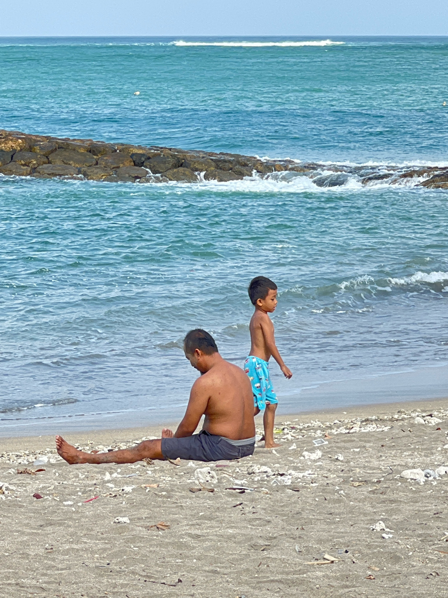 In this photograph, a father and son are enjoying a day at the beach in Kuta, Indonesia. The father, shirtless and wearing dark shorts, sits on the sandy shore with his legs extended. His son, wearing bright blue swim trunks with a patterned design, stands a short distance away, facing the ocean and appearing to be walking along the water's edge.
The scene is set against the backdrop of the vast turquoise ocean, with gentle waves rolling towards the shore. In the distance, a rocky breakwater breaks the rhythm of the waves, creating a calmer area of water. The sky above is a pale blue, suggesting a bright, possibly overcast, day. The beach itself is a mix of sand and scattered debris, likely natural elements like shells and driftwood, along with some man-made litter, indicating it's a well-used, perhaps not perfectly maintained, public beach.
The overall mood appears relaxed and serene, with the father and son enjoying their time together in a natural setting. The focus on the two figures, with the expansive ocean and beach surrounding them, evokes a sense of peaceful coexistence with nature. The image captures a simple moment of family leisure in a tropical coastal environment. There is no visible text in the image.