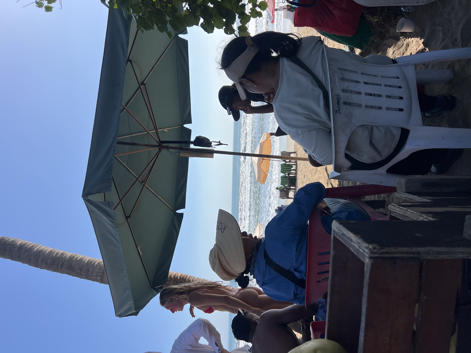 The image depicts a beach scene in Kuta, Indonesia. In the foreground, two individuals are seated under a large green umbrella. One person, viewed from behind, wears a blue shirt and a wide-brimmed hat, while the other, also from behind, is in a light-colored jacket and a visor. To their left, a partially visible person in a white shirt stands near a woman in a red bikini. In the background, the ocean with moderate waves is visible, along with several beach umbrellas and chairs. A palm tree trunk is prominent on the left side of the frame, and the sandy beach extends towards the water. The overall atmosphere suggests a relaxed beach setting on a sunny day.