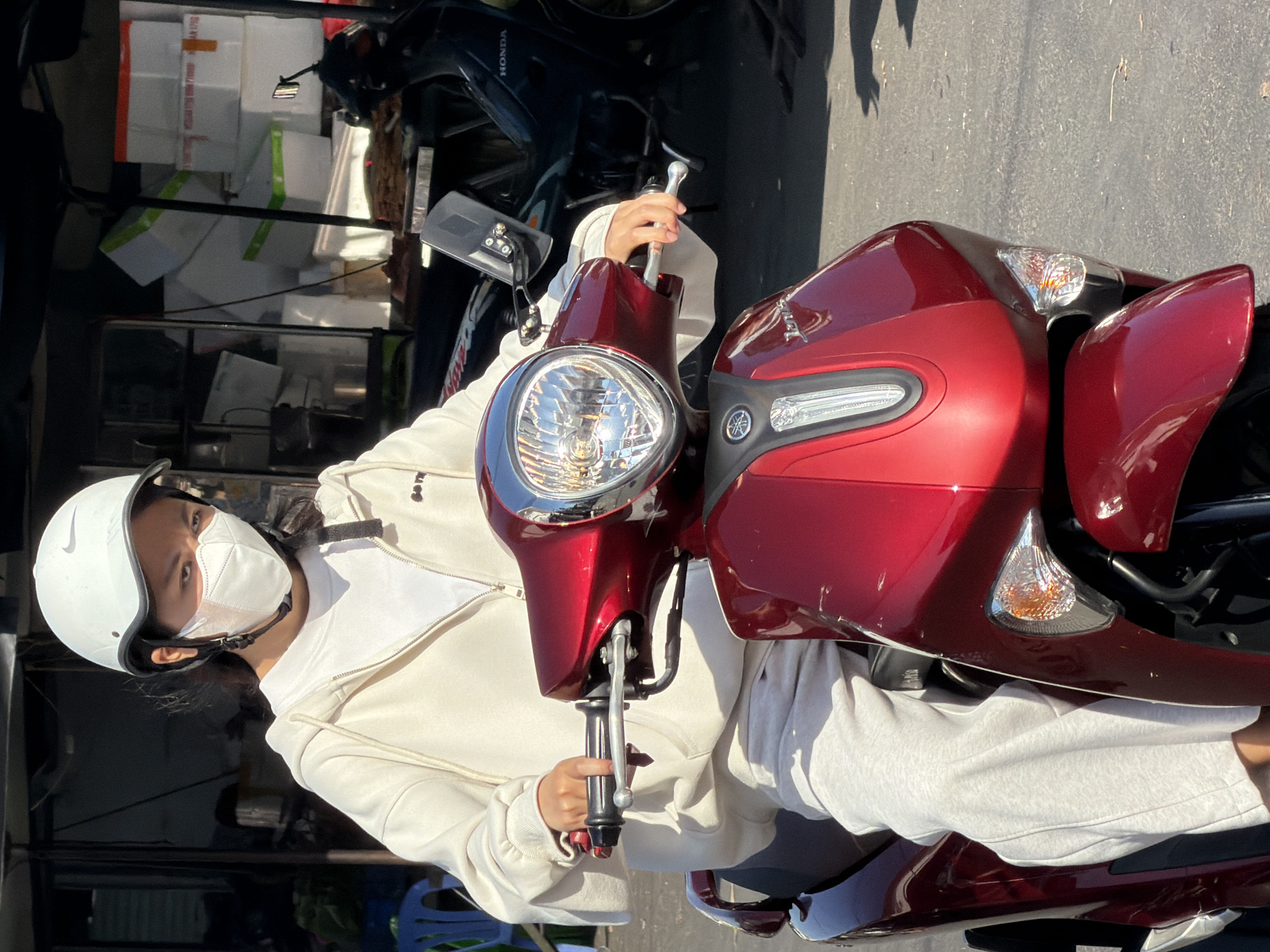 A young woman wearing a white helmet, a white face mask, a white zip-up hoodie, and grey sweatpants is riding a red scooter in Nha Trang, Vietnam. The scooter has a prominent chrome headlight and a Yamaha logo on the front. In the background, there are several other scooters, a dark-colored Honda scooter, and stacks of white boxes with green trim. The scene appears to be in an outdoor urban setting, possibly near a market or shop.