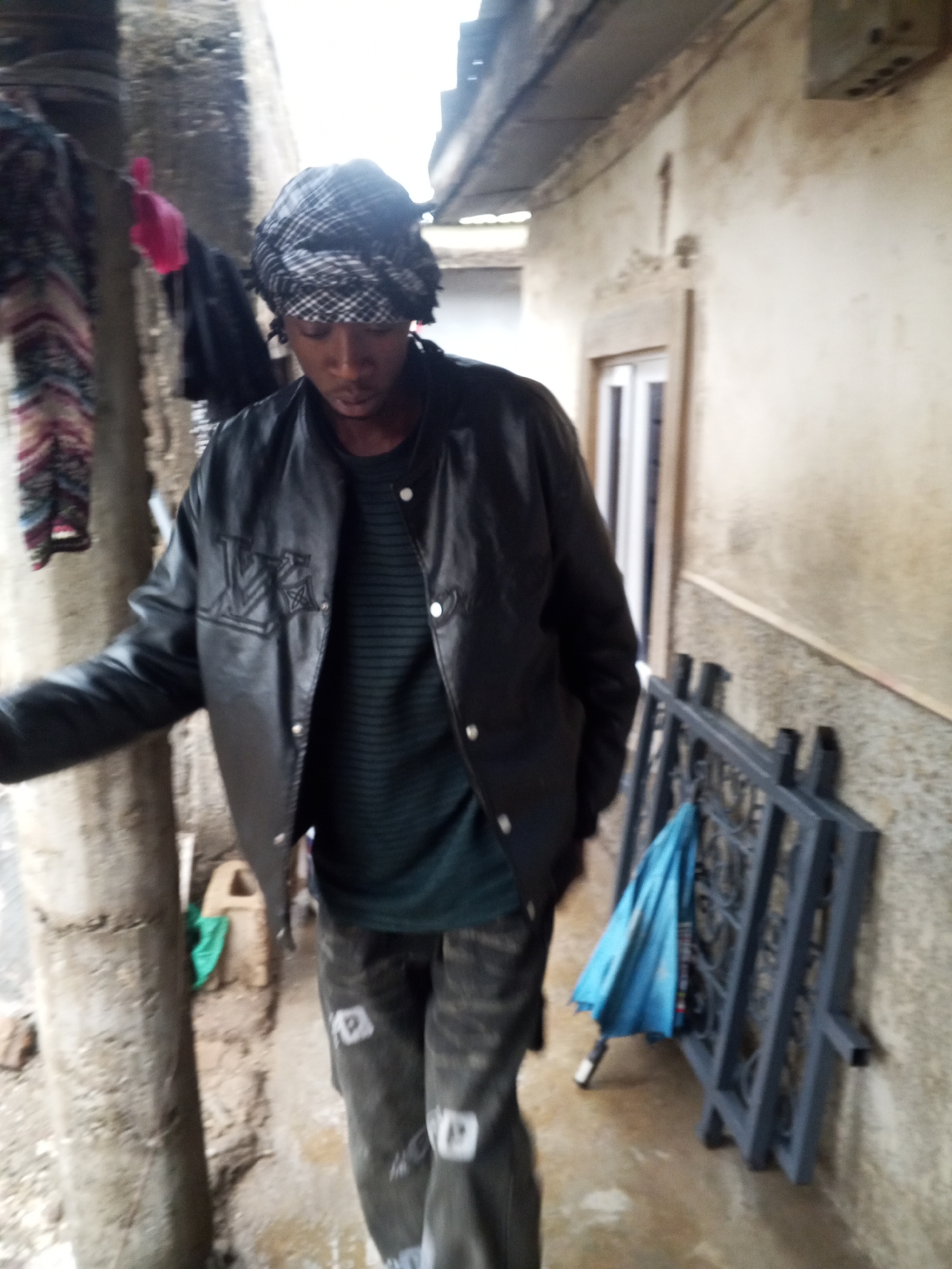 A person is captured in a full-length, slightly blurry shot, walking or standing in a narrow outdoor passage between buildings. The individual, with dark skin, is seen from the waist up, facing slightly left and looking downwards. They wear a black and white checkered head wrap, a black leather-like jacket with an embroidered or appliqued design on the left side (resembling a stylized "LV" monogram), a dark green striped t-shirt, and dark patterned pants.
To the left, a light-colored pillar or post is visible, with patterned fabric and a bright pink garment hanging in the background, suggesting a clothesline. The ground appears wet and potentially muddy around the person's feet.
On the right, a light-colored, weathered building wall features a white-framed window. A folded blue umbrella leans against a dark grey metal gate or rack on the ground. An overhead structure, likely a roof or overhang, is visible above the wall. The diffused lighting suggests an overcast day or a shaded area. The scene depicts an urban or semi-urban environment, with visible wear and tear on the structures. No specific activity beyond walking or standing is discernible, and the person's expression is not clearly visible.