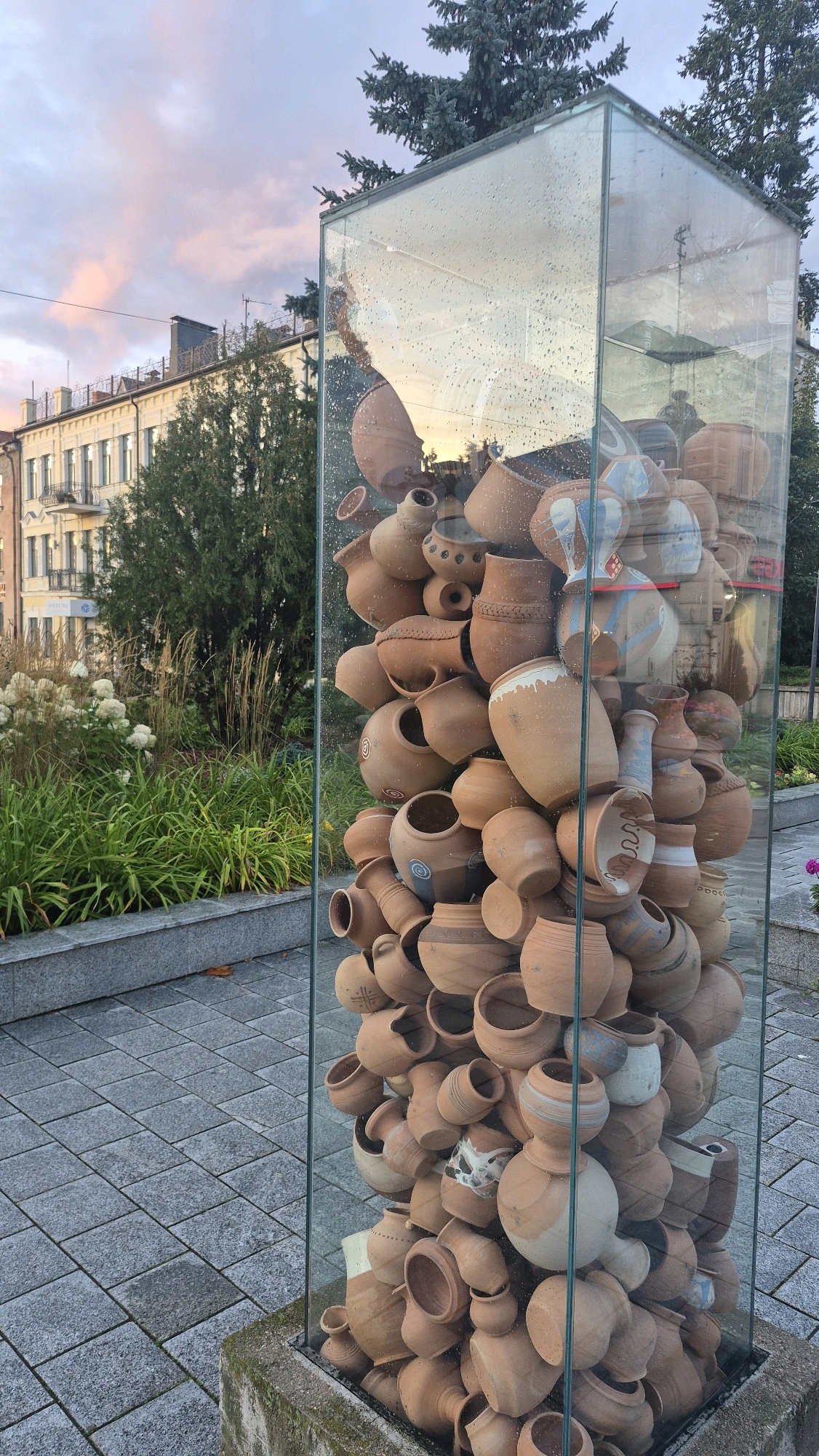 The image displays an outdoor art installation in Daugavpils, Latvia. The central subject is a tall, rectangular glass case filled with numerous terracotta pottery items of various sizes and shapes. The pottery is densely packed, with many pieces overlapping and leaning against each other. Some of the pottery features decorative patterns, including what appear to be swirling blue designs and geometric motifs.
The glass case is slightly wet, suggesting recent rain, with numerous water droplets visible on its surface, creating reflections of the surroundings. The installation sits on a concrete base.
In the background, a building with a light-colored facade and balconies is visible, along with several trees, including evergreen conifers. The sky above is partly cloudy with soft hues of pink and orange, indicative of either sunrise or sunset. In the foreground, a paved area with square stone tiles leads up to the installation. Green foliage and plants are present to the left of the structure. No people are visible in the image.