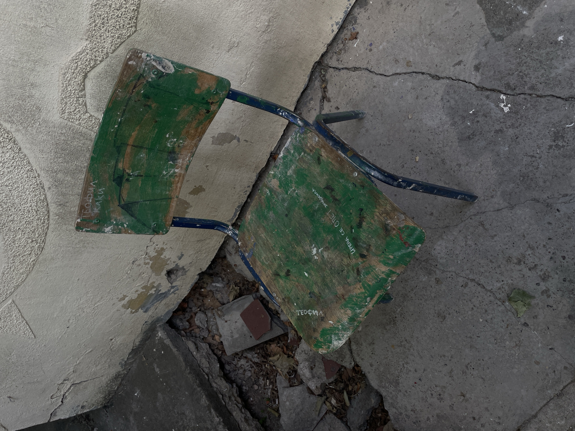 An overhead shot captures a broken, dilapidated chair lying at an angle on a concrete surface, nestled against a textured, light-colored wall. The chair's frame is blue metal, and its backrest and seat are made of wood painted green, with significant wear and exposed wood showing through.
The scene depicts a neglected outdoor area. The concrete ground is cracked and uneven, with dirt, small stones, and dry leaves visible in the crevices and gaps. The wall, likely stucco or rough plaster, also shows signs of wear and damage, with some areas of discolored peeling paint.
No activity is taking place; the chair appears abandoned or discarded. The natural light suggests it is daytime. The overall impression is one of urban decay or a neglected public space in Belgrade, Serbia.
Several texts are visible, written in white or light-colored Cyrillic script on the green surfaces of the chair:
- On the left wooden piece (backrest): "БОДИЛА КАКИ" (BODILA KAKI)
- On the right wooden piece (seat): "ИРЕНА СЕ ОПЕТ" (IRENA SE OPET, translating to "Irena again" or "Irena once more")
- Below "ИРЕНА СЕ ОПЕТ" on the seat: "ТЕОФИЛ" (TEOFIL)