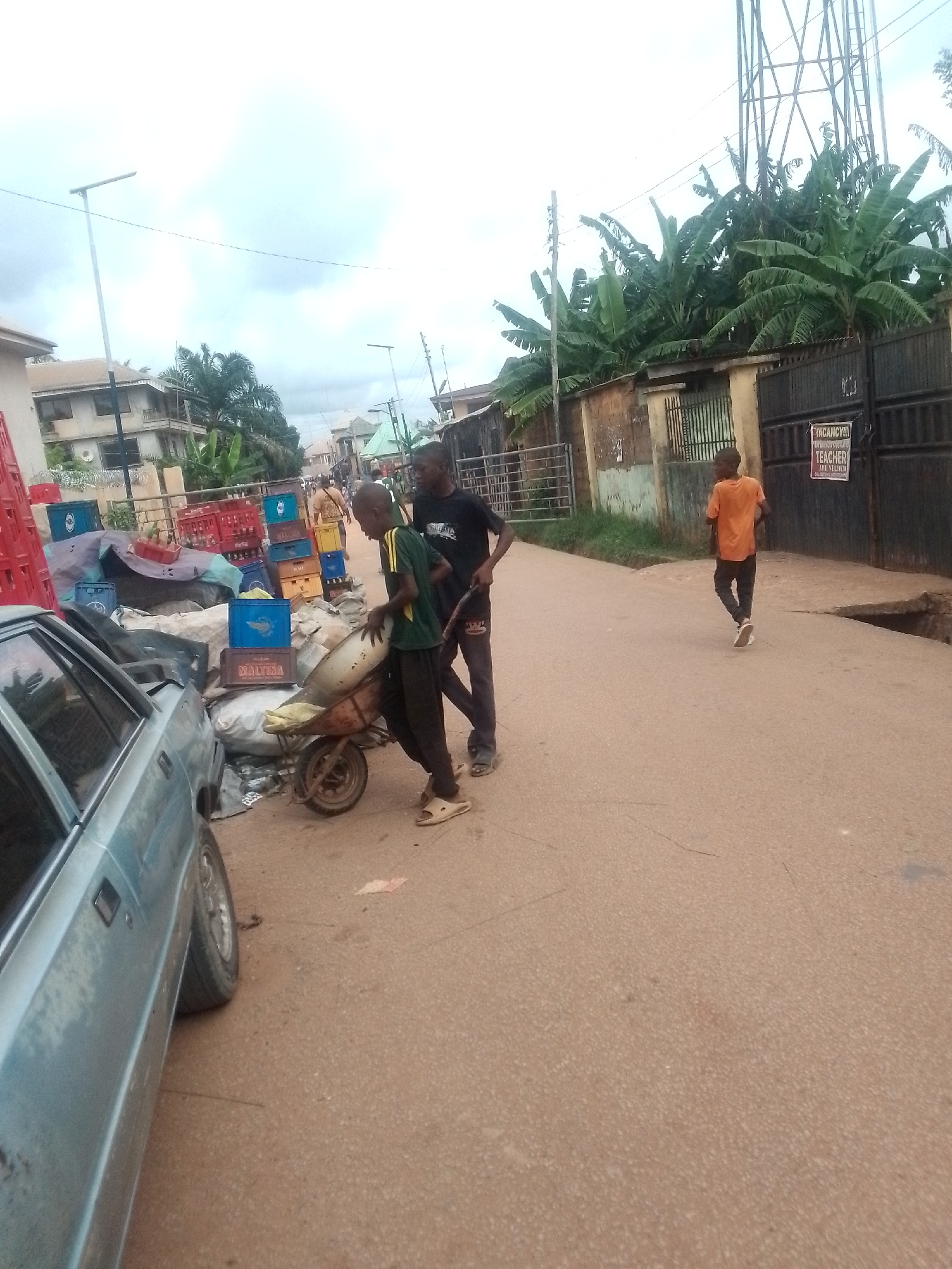 The image depicts a street scene in the city of Unknown, Unknown. Two young men are maneuvering a wheelbarrow filled with materials on a paved road. One man is actively pushing the wheelbarrow, while the other appears to be assisting. To the left, a weathered, light blue car is parked. Adjacent to the car, there is a collection of crates and covered objects. In the background, a person in an orange shirt is walking away down the street. On the right side of the image, there is a gated property with a sign indicating a teaching vacancy. Overhead, power lines and a communication tower are visible against a cloudy sky. The street appears to be in a residential area, with buildings and foliage visible in the background.