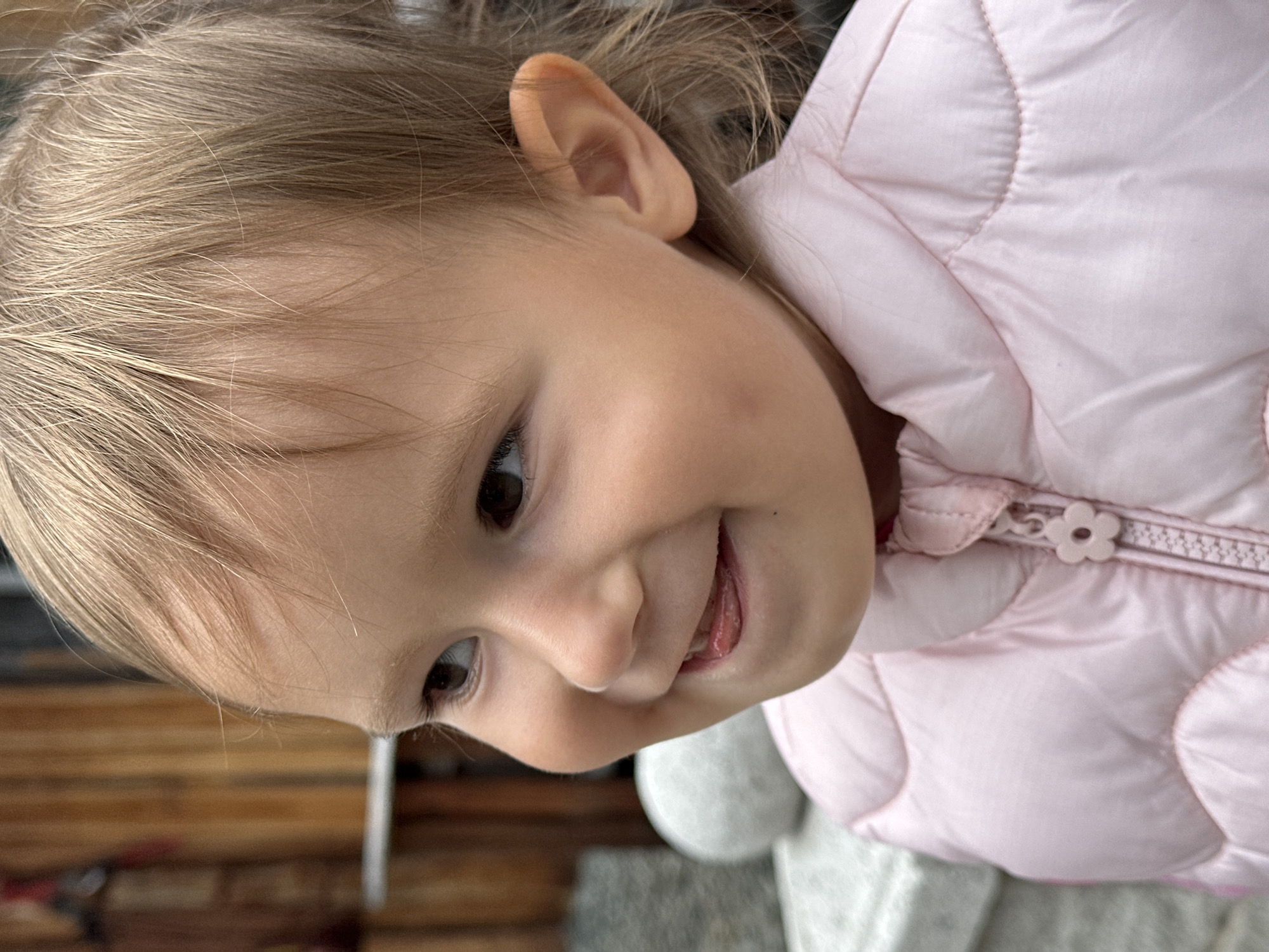 A close-up, profile shot captures a young child with light brown hair and rosy cheeks, wearing a pink puffer jacket. The child is smiling, with their tongue slightly visible. The background consists of wooden planks and a blurred gray sphere. The image is undated and unlocated.