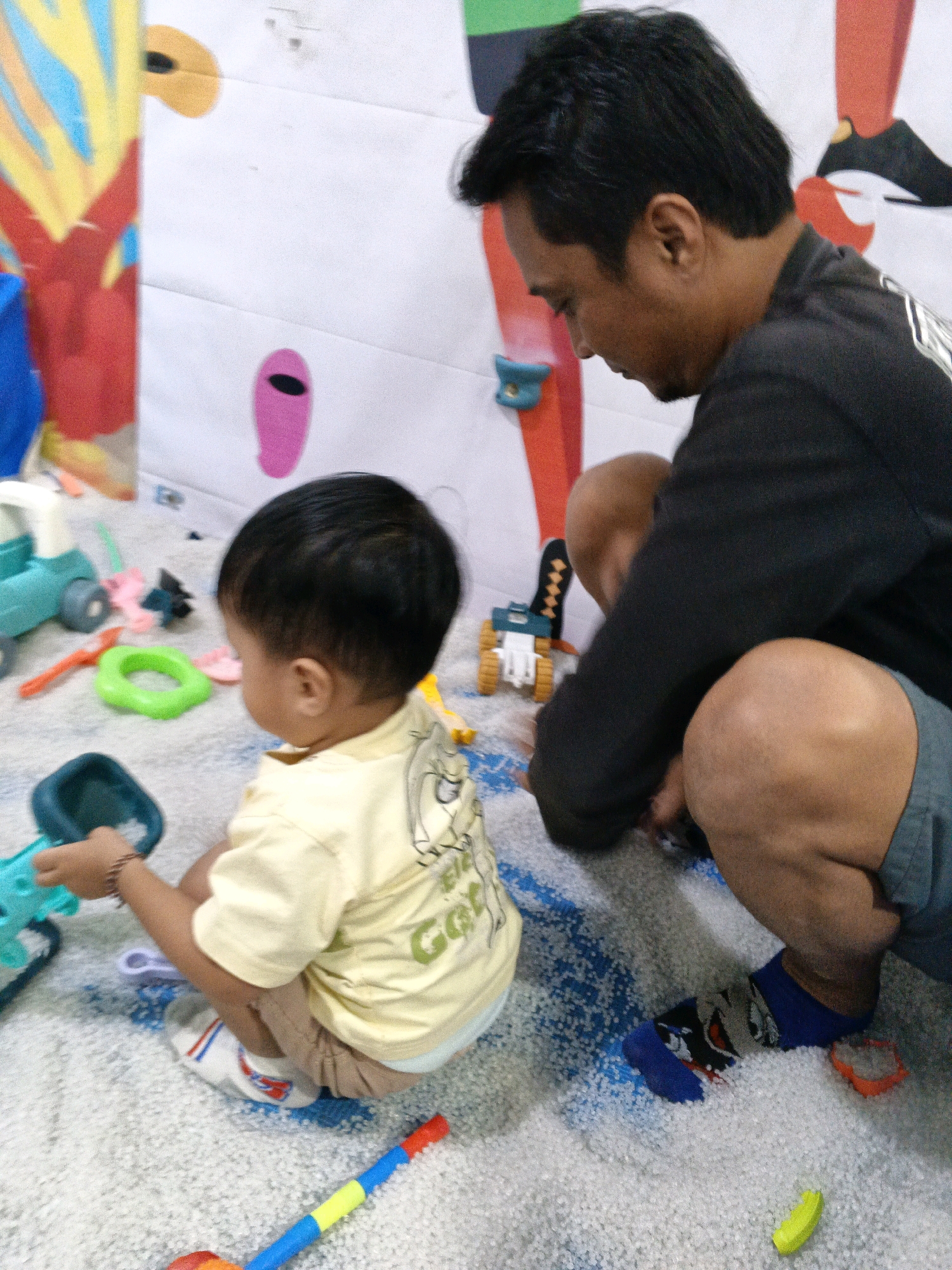 In Blahbatuh, Indonesia, a father and his young child are playing together on a light-colored, textured floor, possibly in a dedicated play area. The father, wearing a black long-sleeved shirt and gray shorts, is crouched down, facing away from the camera and towards the child. He has short dark hair and is looking downwards, engaged in the activity. His right leg is bent, and he is wearing a blue sock with a character print.
The child, seen from behind, is wearing a light yellow short-sleeved shirt with a graphic on the back and light brown shorts. They are also crouched, facing the father and the toys. The child is holding a blue plastic toy scoop.
Scattered around them are various colorful plastic toys, including a teal and white toy vehicle, a green ring, and a red and blue striped object resembling a wand or stick. The background appears to be a white wall with colorful abstract shapes and depictions of legs and feet. The overall environment suggests a casual, playful setting for the father and child's interaction.