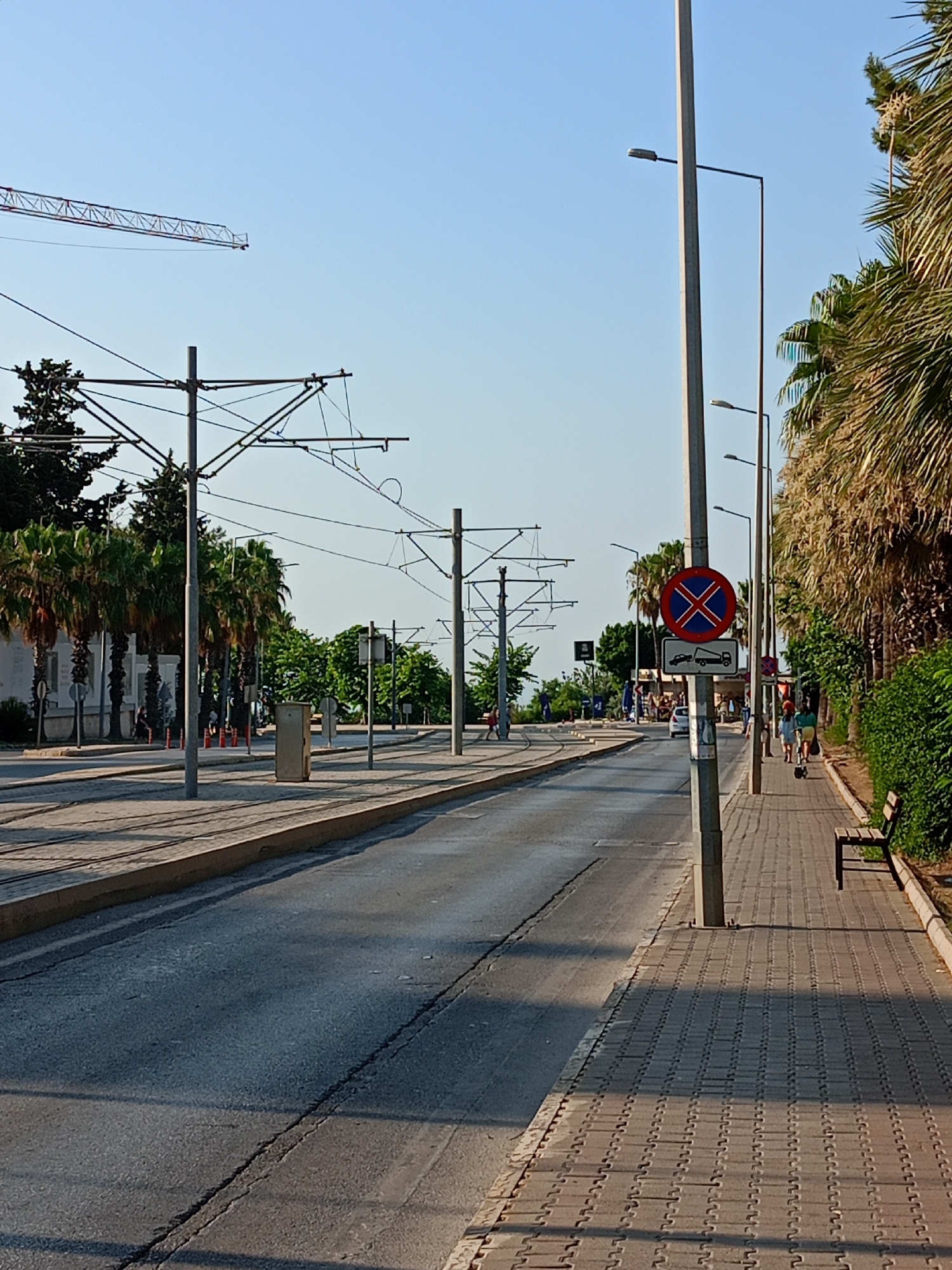 The image captures a scene on a sunlit street in Antalya, Turkey, likely along a tram line. The foreground shows a paved road with tram tracks embedded. To the right of the road, a sidewalk is paved with rectangular blocks. Along the sidewalk, a wooden bench is visible, backed by dense green foliage and tall palm trees. A prominent lamppost stands on the sidewalk, bearing a circular red and blue traffic sign with a white "X" and an additional rectangular sign below it depicting a tow truck. In the background, more palm trees and various buildings can be seen. The sky is clear and blue. Several overhead wires for the tram system are suspended by poles on either side of the road. A construction crane is visible in the far distance. A few individuals are present, walking on the sidewalk, their actions and interactions not clearly discernible. The overall impression is one of an urban environment in a warm climate.