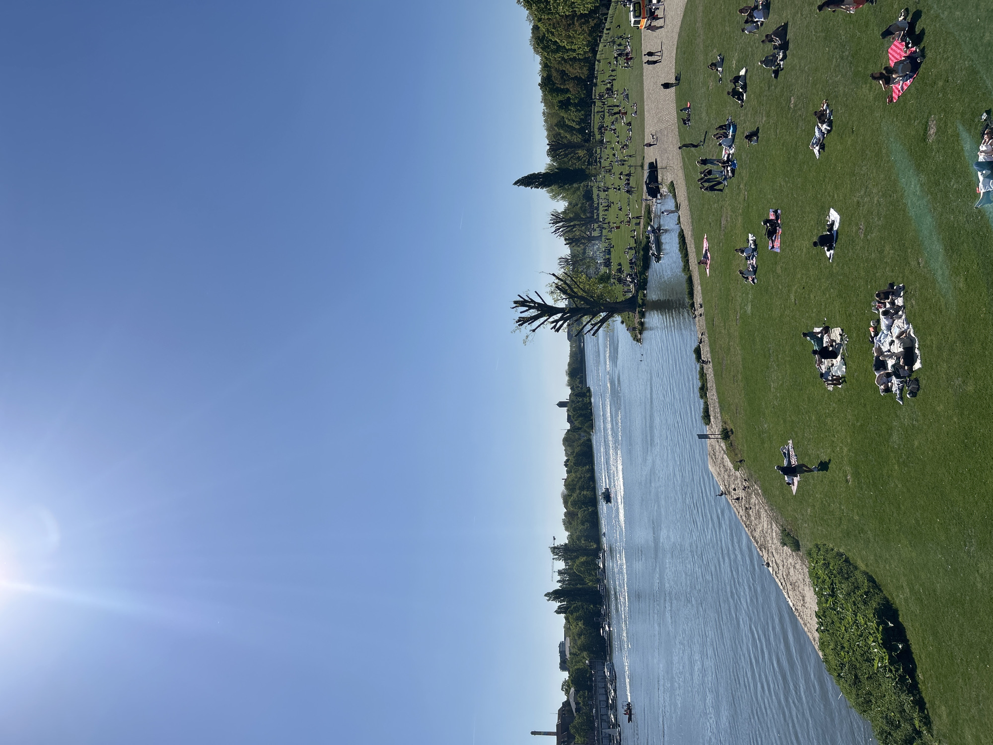 A bright, sunny day shows a wide river and a green park with numerous people relaxing on the grass. The river stretches towards the horizon, reflecting sunlight. Along the riverbank, people are spread out, some lying on blankets, others in small groups. Trees line the edge of the park. The scene suggests a relaxed, recreational environment. The location appears to be an urban park along a riverfront.