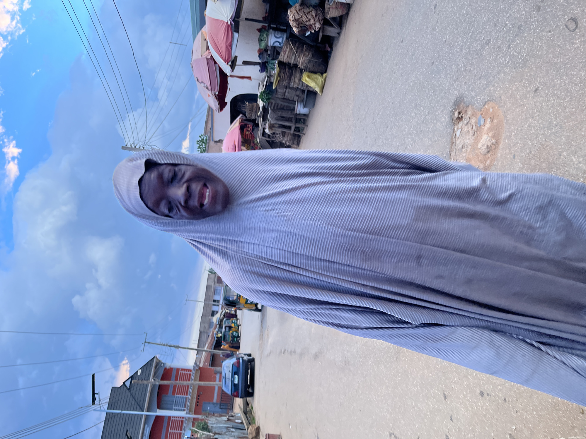The image captures a lively outdoor scene, featuring a person smiling broadly in what appears to be a street or market setting during the day. The main subject is a person, likely a woman, with dark skin, wearing a light grey or light blue full-body garment that covers her head and drapes loosely over her body, characterized by a pleated texture. Her face is visible, and she is expressing joy with a wide, open-mouthed smile or laugh, showing her teeth.
The background reveals a bustling environment. To the person's right (when oriented correctly), several market stalls are visible, some covered by large, colorful umbrellas (pink/red and white), under which various goods like woven baskets and sacks are stacked. Further down the street, multiple vehicles, including cars and auto-rickshaws (tricycles), are present, indicating a busy thoroughfare. On the opposite side of the street, there are buildings, some with reddish-brown facades and others with grey roofs, along with utility poles and overhead power lines. The sky above is bright blue with scattered white clouds, suggesting clear, sunny weather. The ground is a paved road surface, appearing to be asphalt or concrete, with some visible wear and patches of dirt. The overall scene suggests daily life in an urban or semi-urban area, possibly in a West African region given the market setup and types of vehicles. No discernible text is visible in the image.