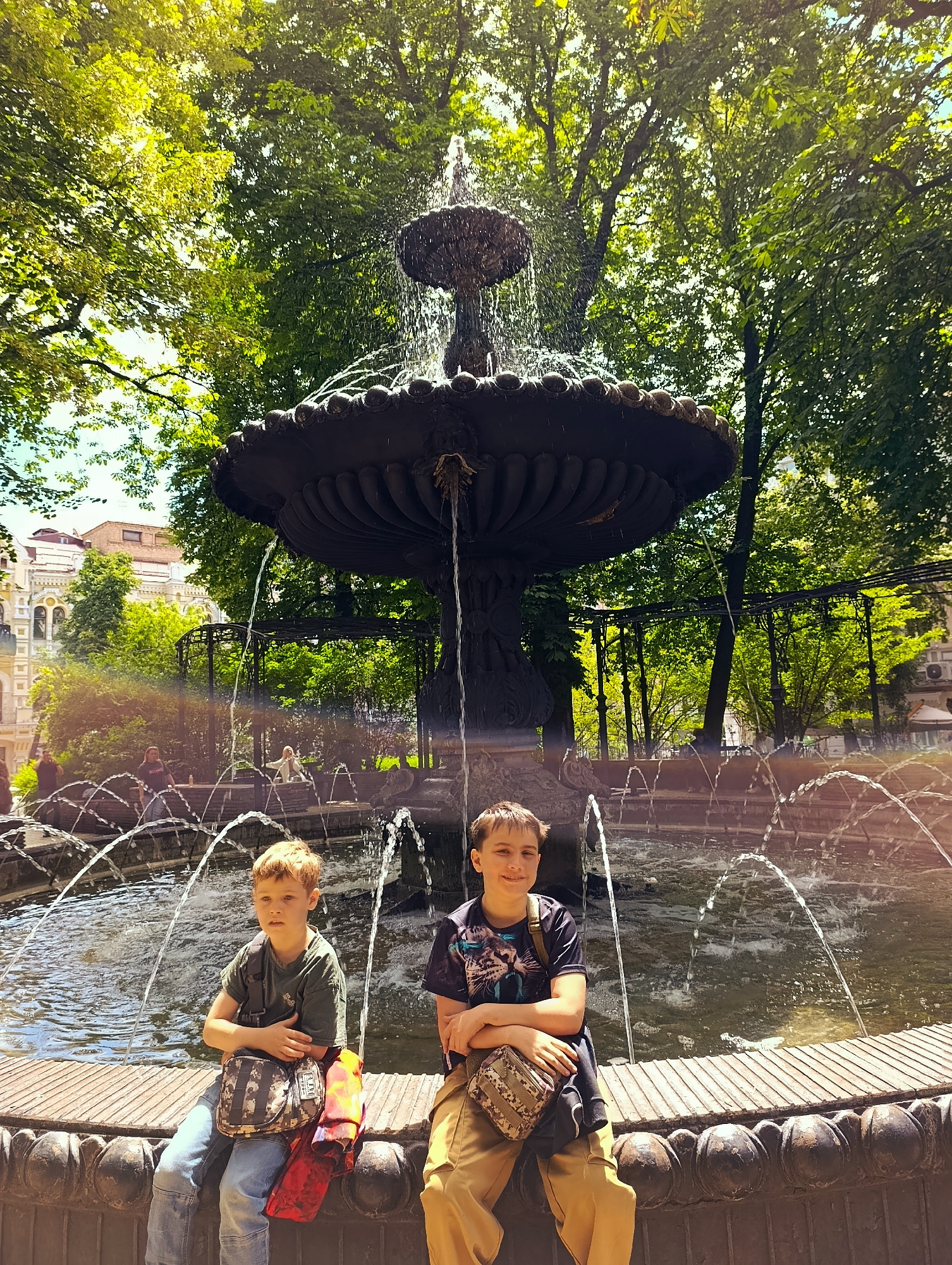Two young boys are seated on the edge of a decorative fountain in a park. The boy on the left, with light brown hair, wears a green t-shirt, blue jeans, and a camouflage-patterned crossbody bag. A red and orange item, possibly a jacket, is slung over his left leg. The boy on the right, with darker brown hair, is wearing a dark t-shirt with a graphic design and light brown pants, also with a camouflage-patterned crossbody bag. The fountain has multiple tiers and sprays water upwards, with jets also arcing over the pool. Lush green trees form a canopy overhead, dappling the scene with sunlight. In the background, an ornate building with balconies and a brick facade is visible, suggesting an urban park setting, likely in Kyiv, Ukraine, given the context. The overall atmosphere is bright and sunny.